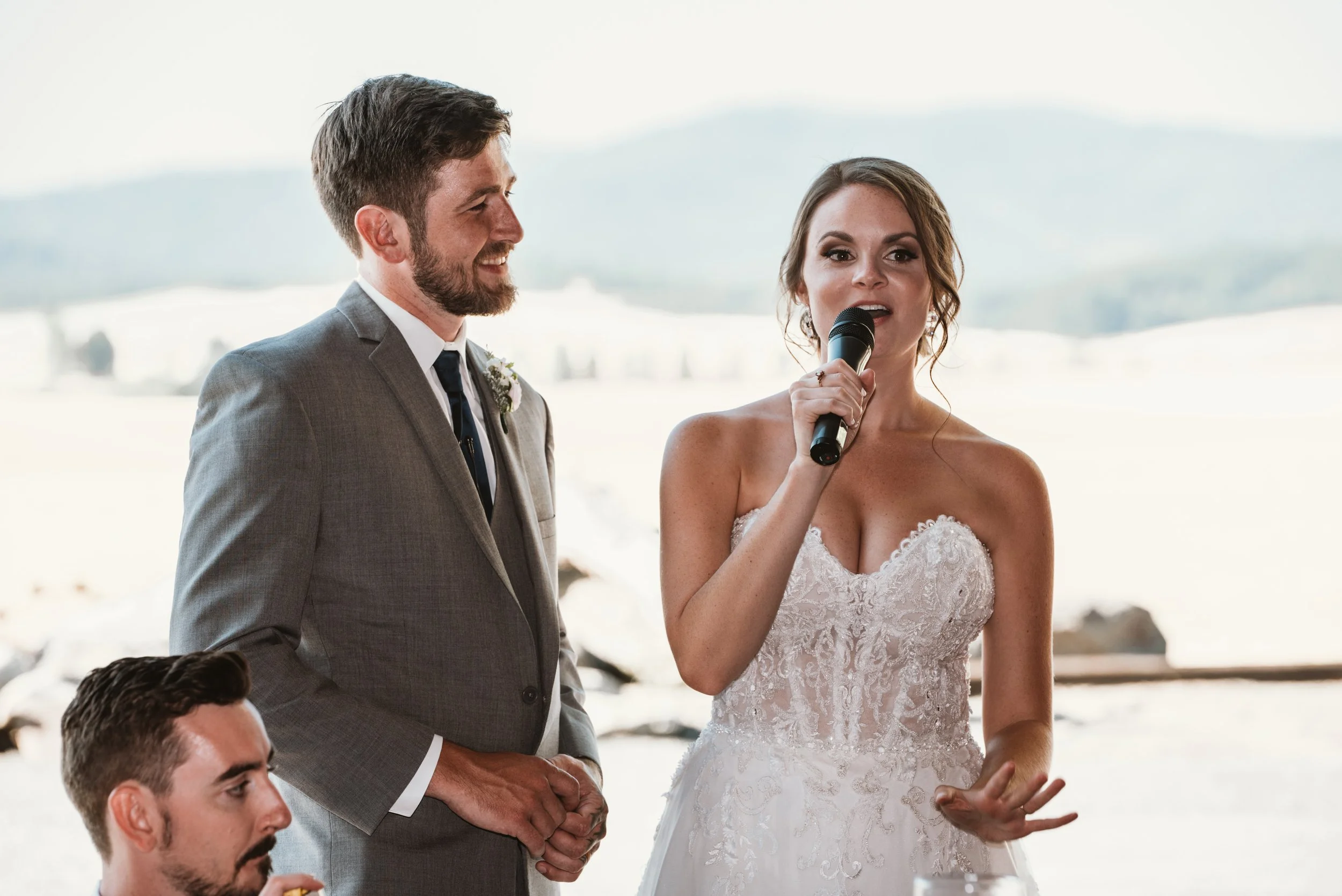 Bride and groom at wedding reception, bride speaking into microphone, groom smiling, mountainous background.