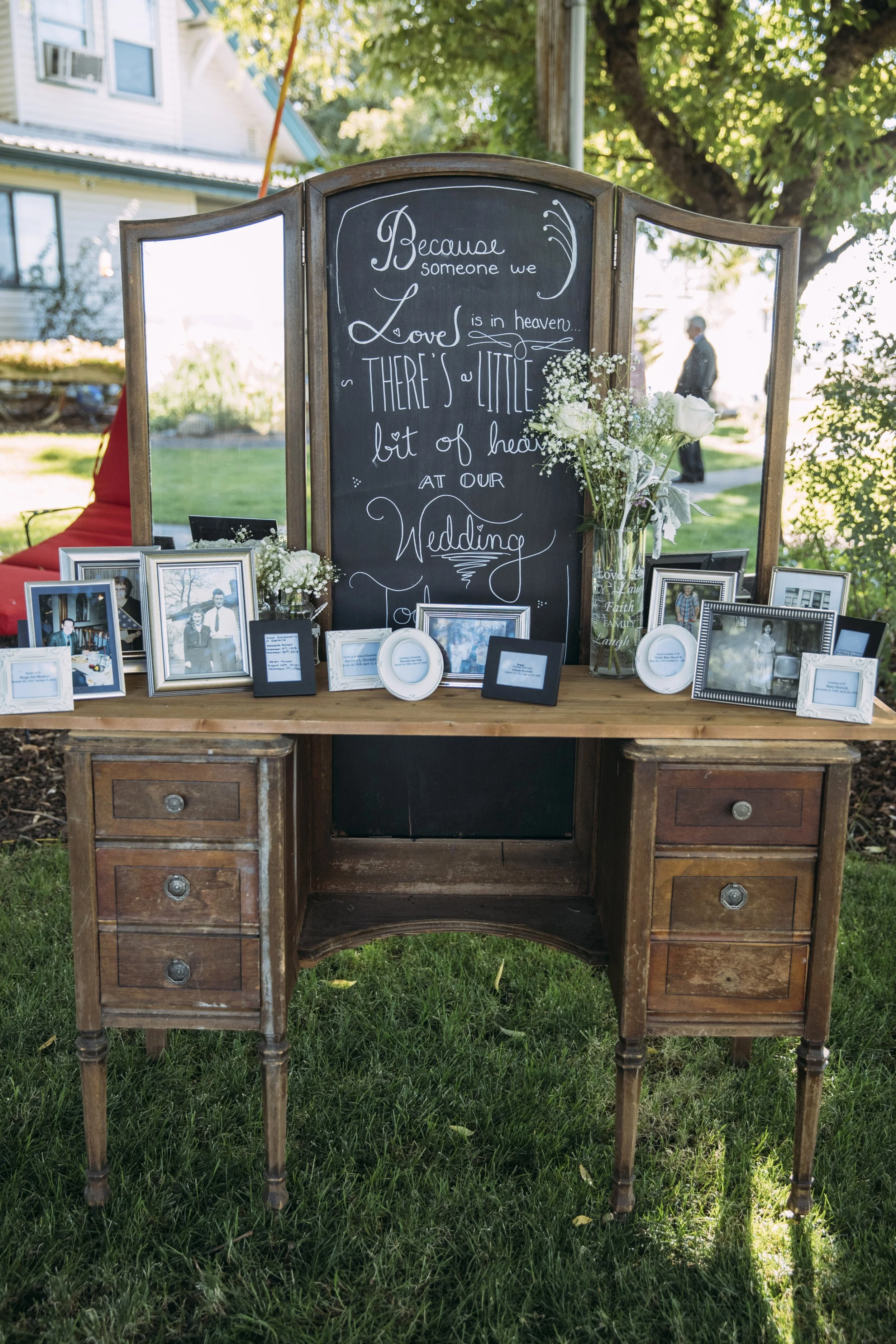 Wedding memorial table with framed photos, a chalkboard sign saying “Because someone we love is in heaven, there’s a little bit of heaven at our wedding,” and flowers, set on a wooden dresser in an outdoor setting.