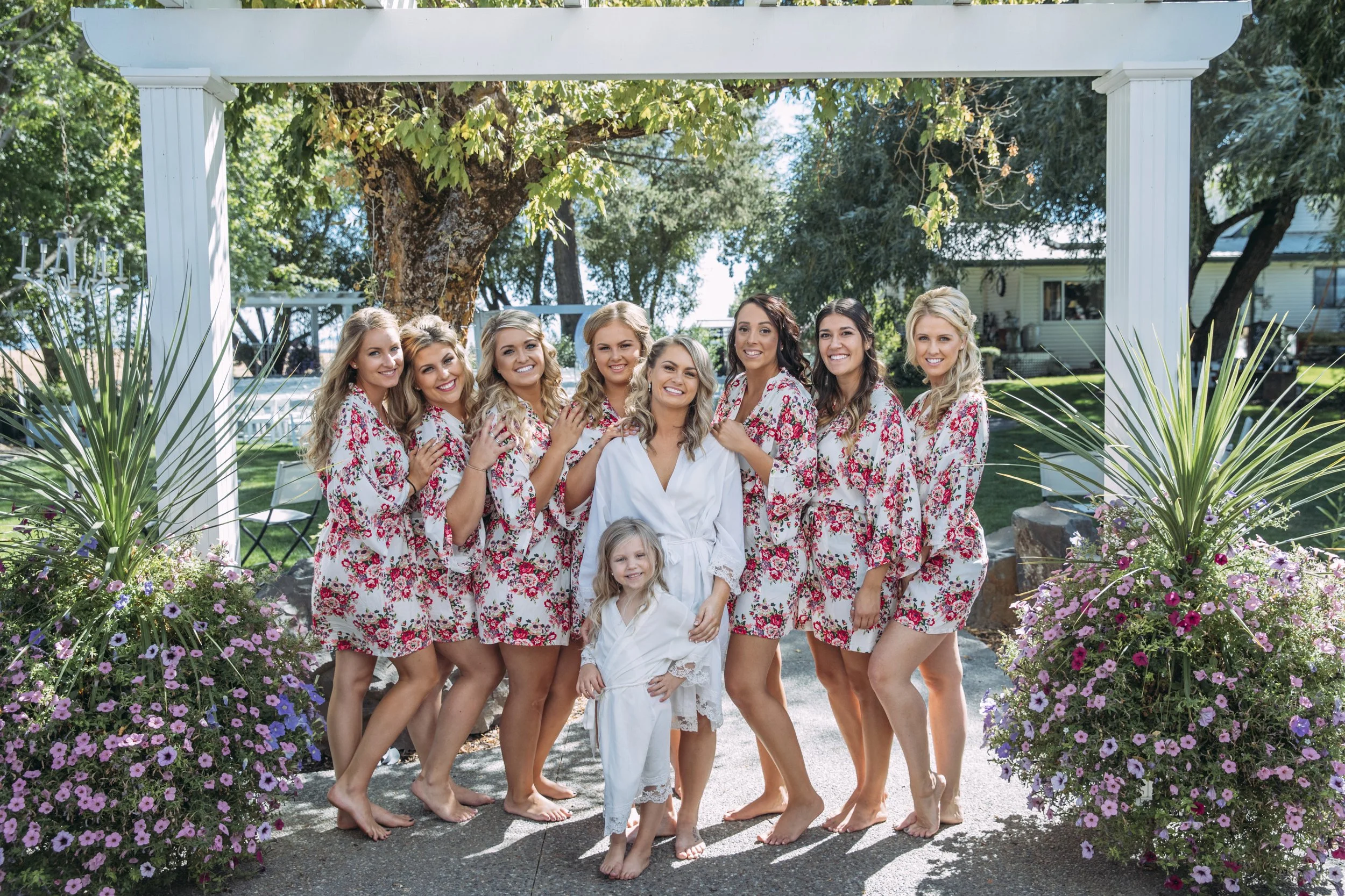 Group of women and a child wearing matching floral robes, posing outdoors under a pergola with plants and a tree in the background.