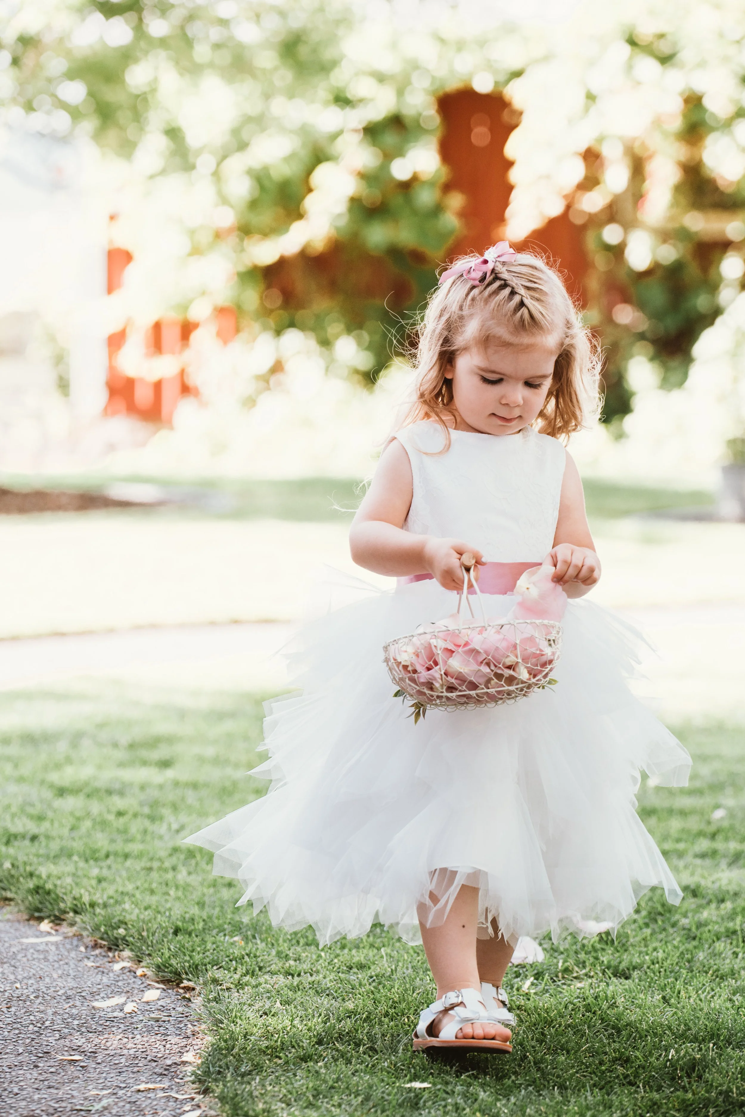Young girl in a white dress holding a basket of pink flowers on a grassy path.
