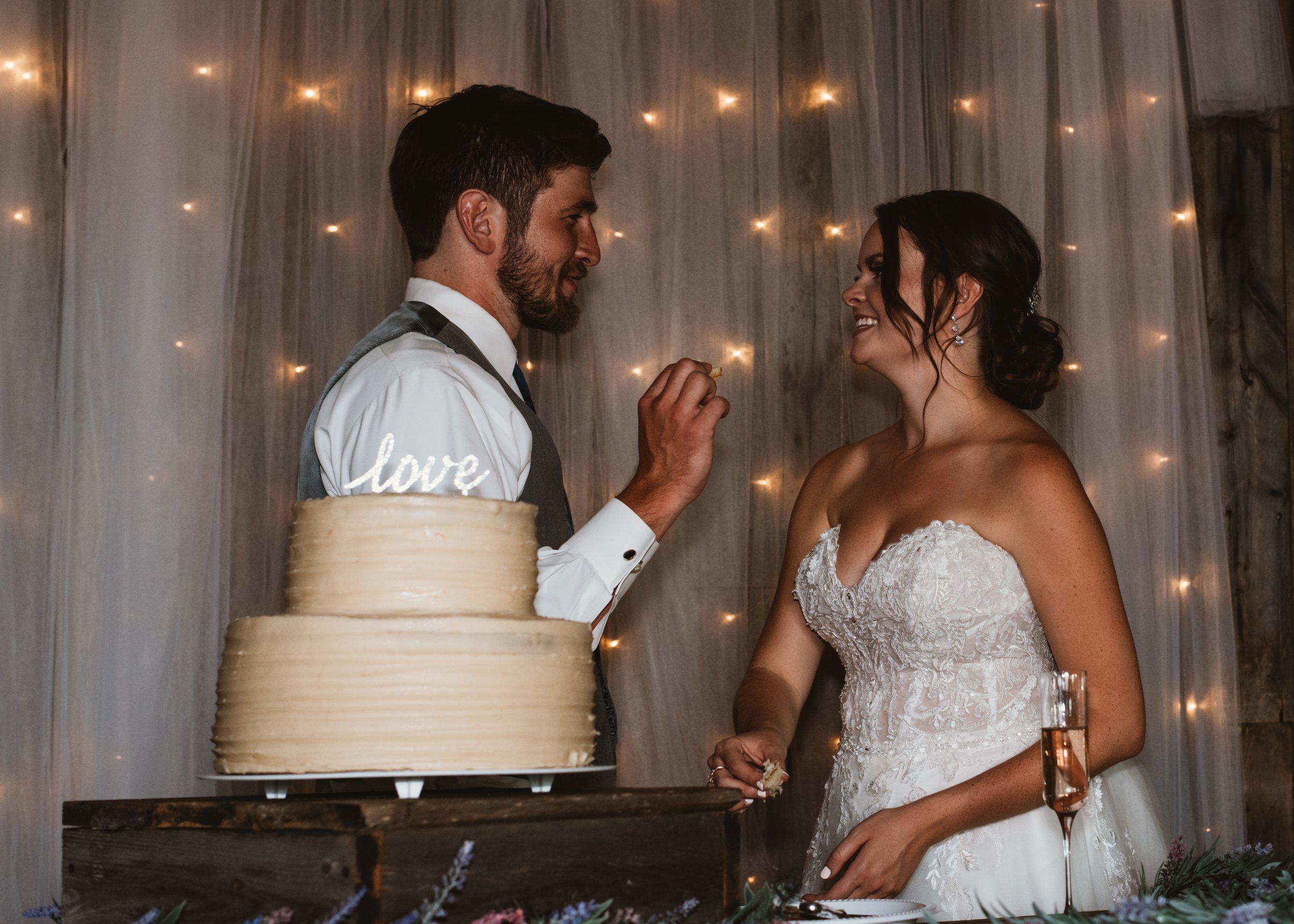A bride and groom smiling at each other while feeding cake. The cake is on a stand with a "love" topper, and the background features string lights.