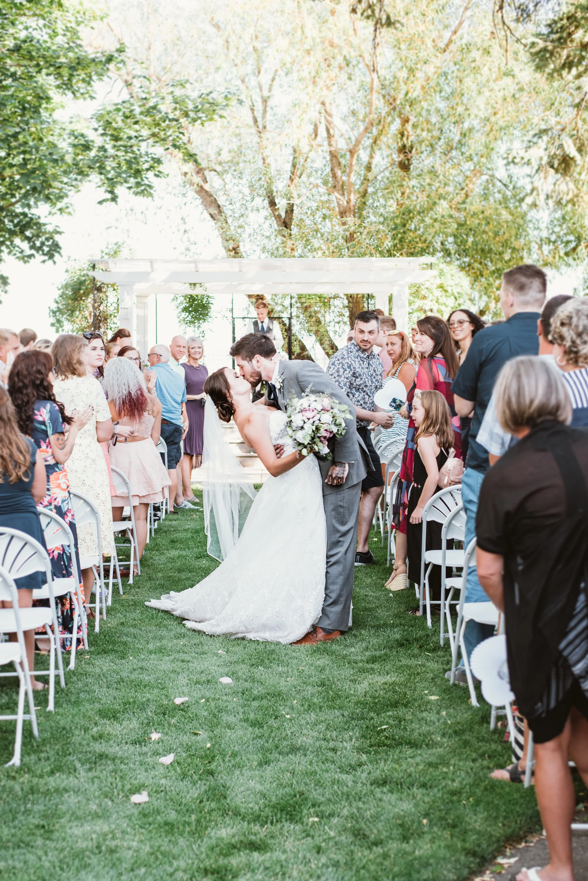A newlywed couple kissing at an outdoor wedding ceremony, surrounded by seated guests on either side on a grassy aisle, with a white arch in the background.