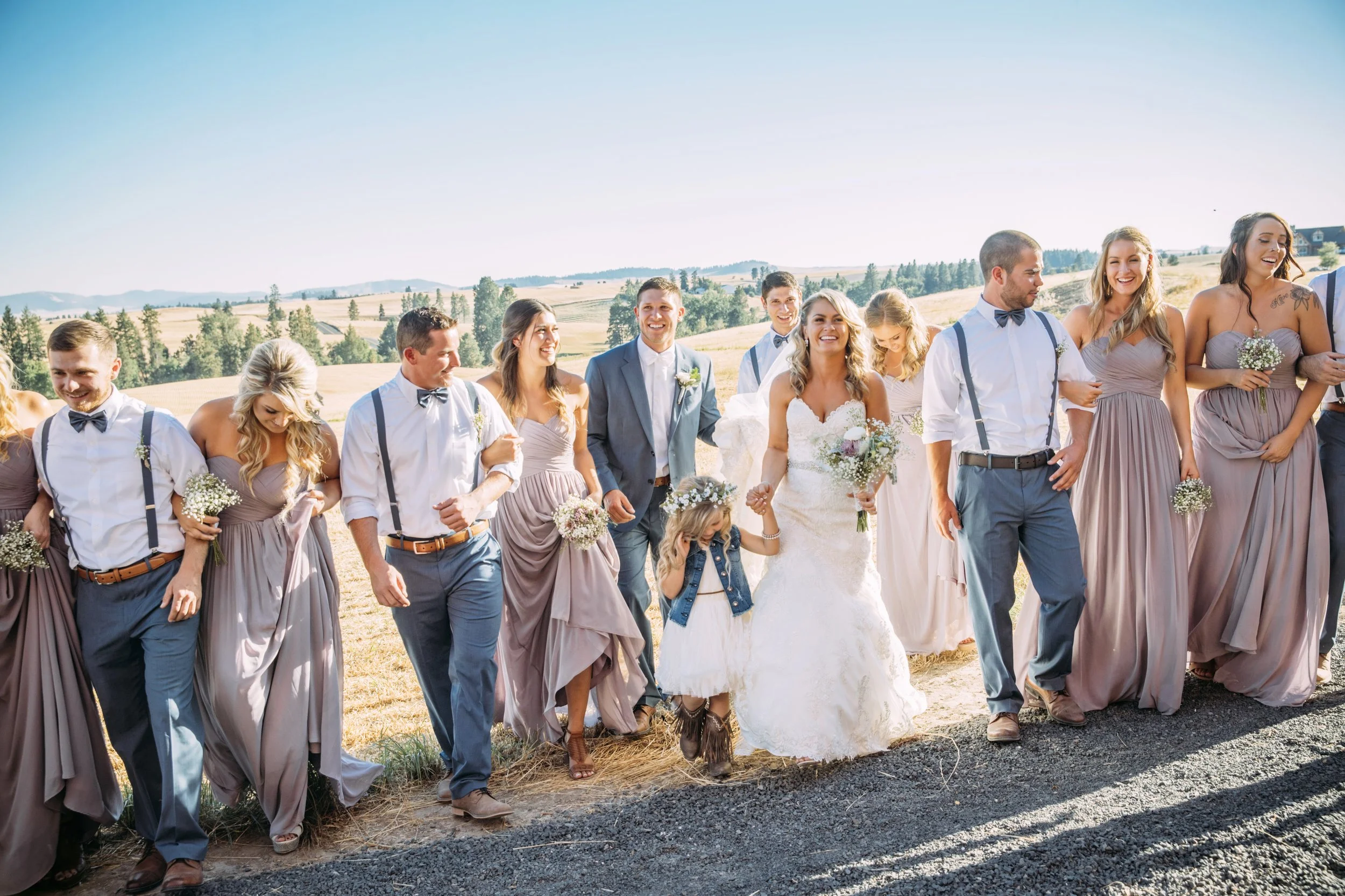A wedding party walks together outdoors, consisting of bridesmaids in lavender dresses, groomsmen in gray pants with suspenders, and a bride in a white gown. The setting is a rural landscape with fields and trees visible in the background.