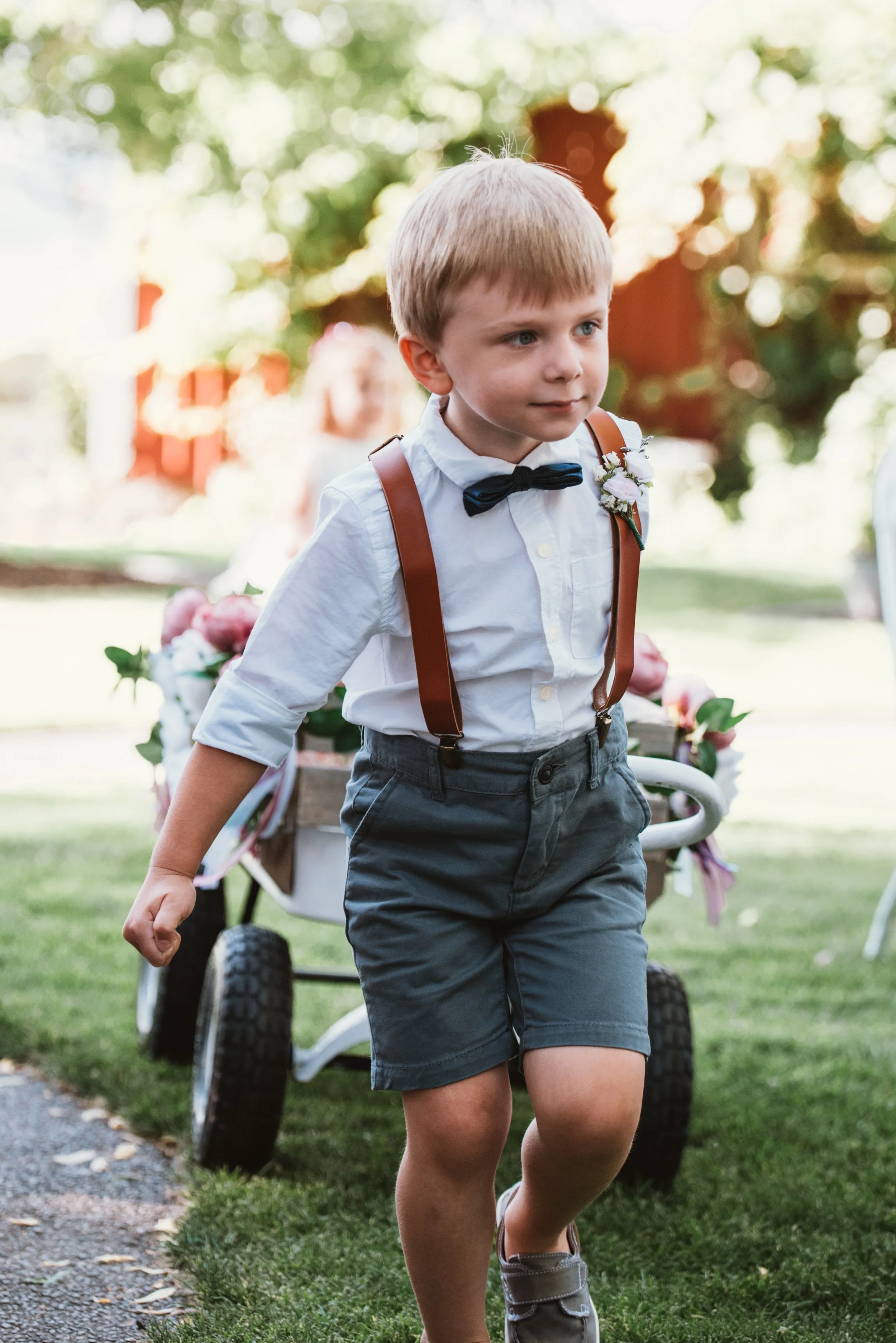 Young boy at a wedding wearing a white shirt, bow tie, and suspenders, pulling a flower-decorated cart.