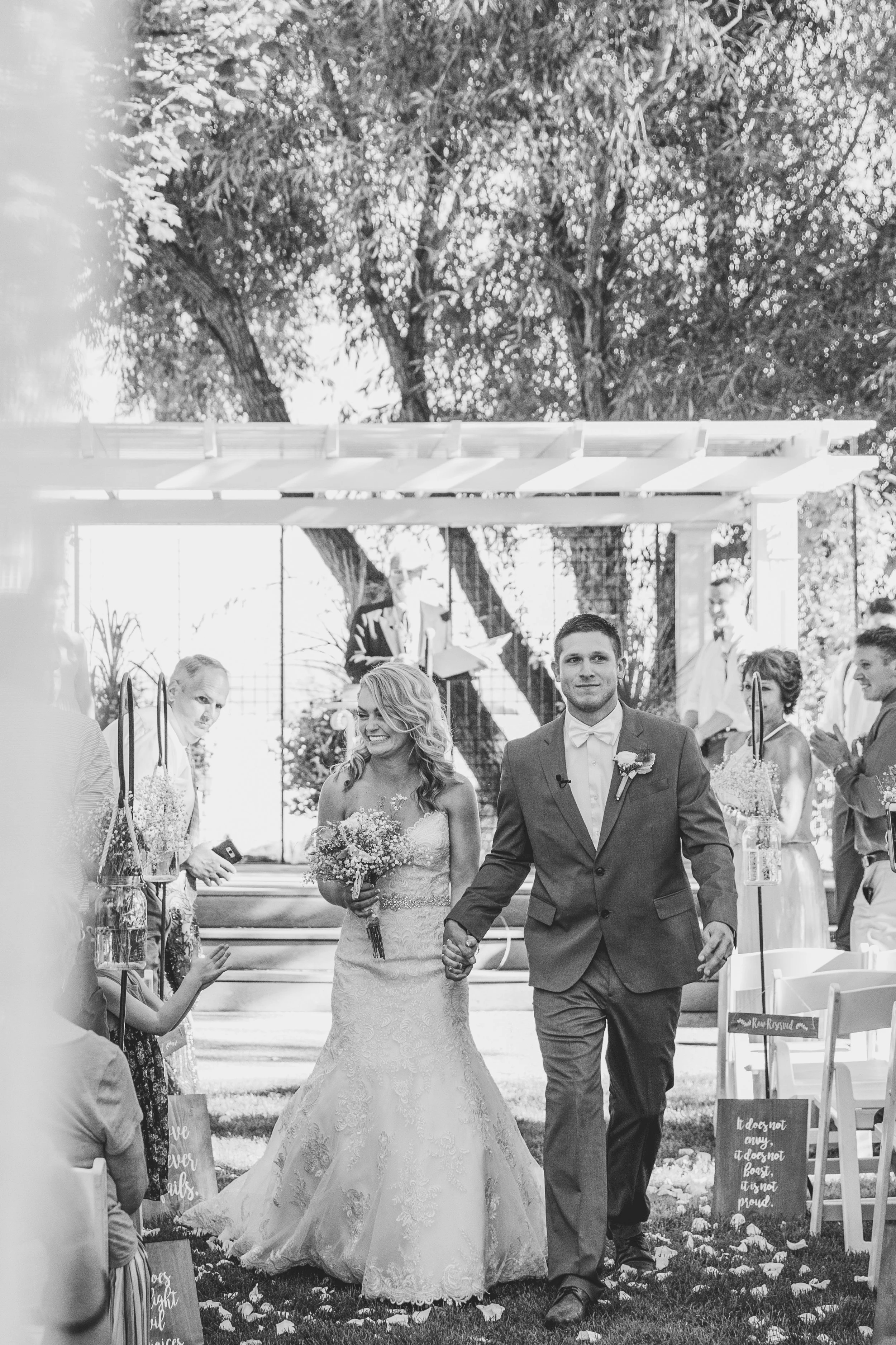Bride and groom walking down the aisle during an outdoor wedding ceremony, surrounded by guests. Black and white photo.