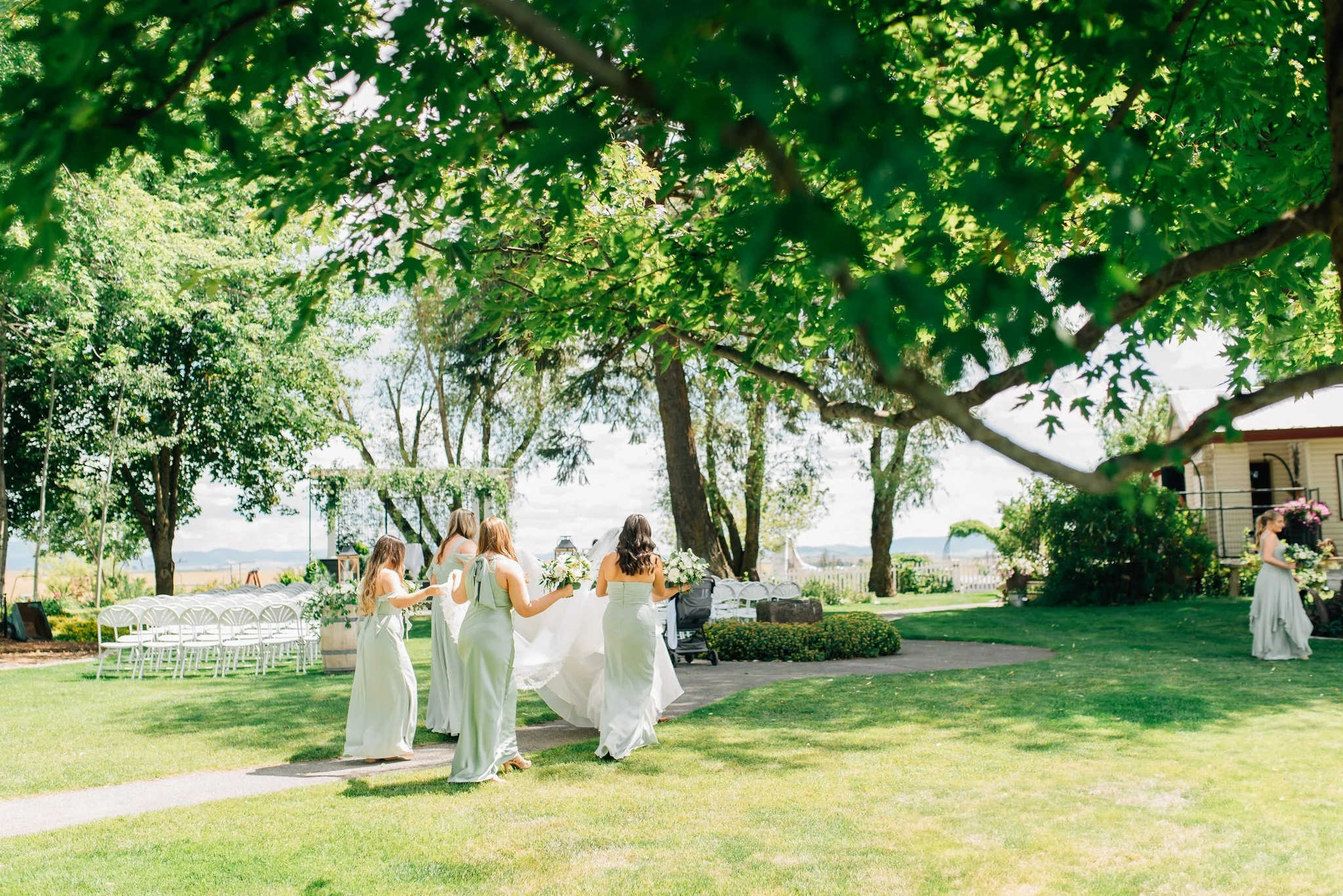 A group of women dressed in light green dresses, holding bouquets, walking outdoors on grass under large green-leafed trees in a park or garden setting, with chairs arranged in the background, possibly at a wedding or special event.