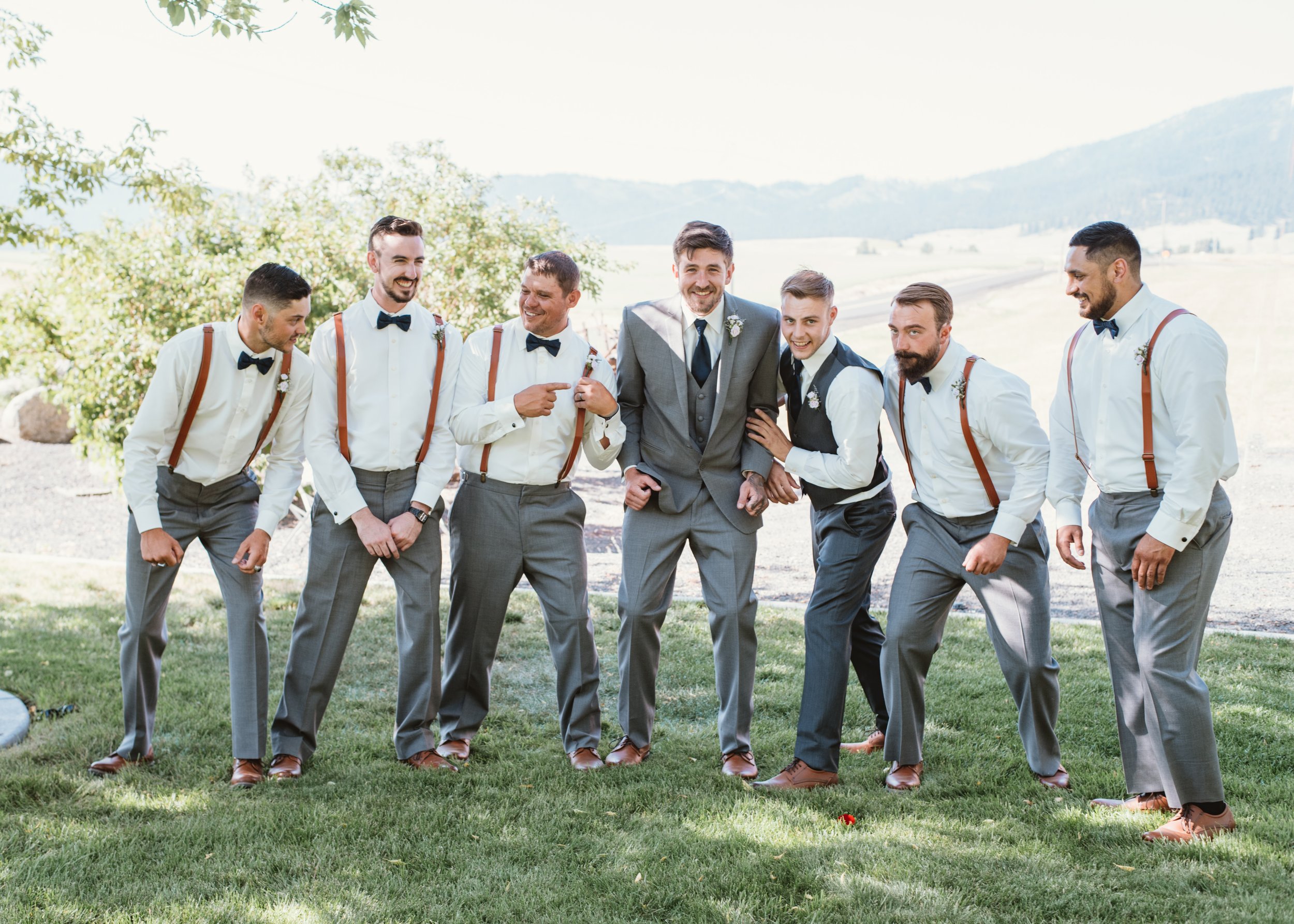 Group of seven men wearing formal attire, including suspenders and bow ties, standing and smiling in a grassy outdoor setting.
