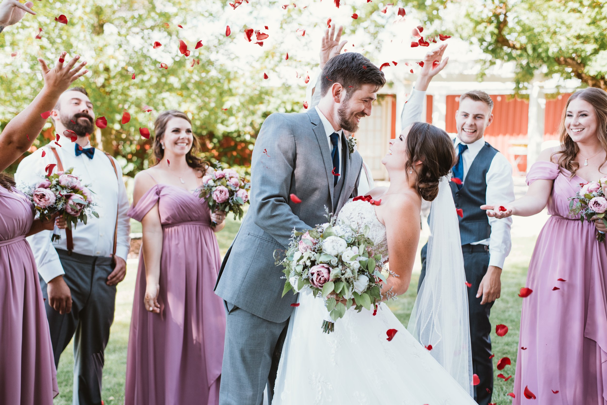 Wedding celebration with a bride and groom surrounded by bridesmaids in purple dresses and groomsmen in formal attire, throwing red rose petals.