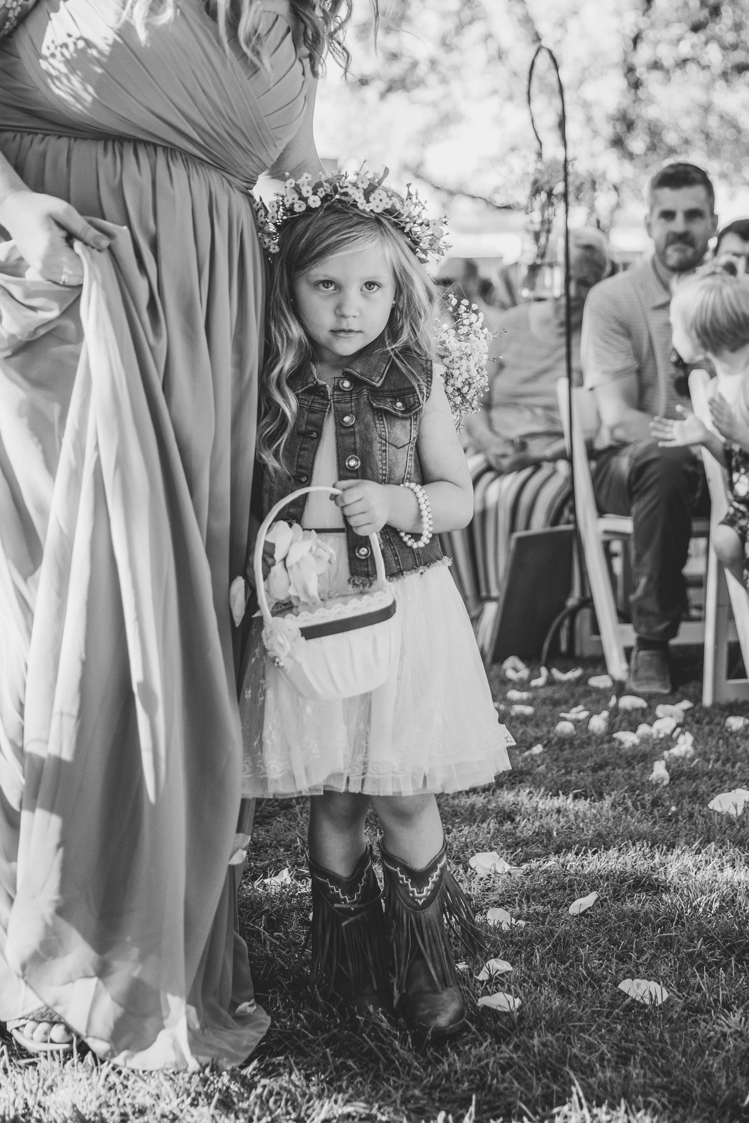 A black and white photo of a young girl at a wedding, wearing a flower crown, a denim vest over a white dress, holding a basket. She is standing beside a woman in a gown. There are people seated in the background and flower petals scattered on the gr