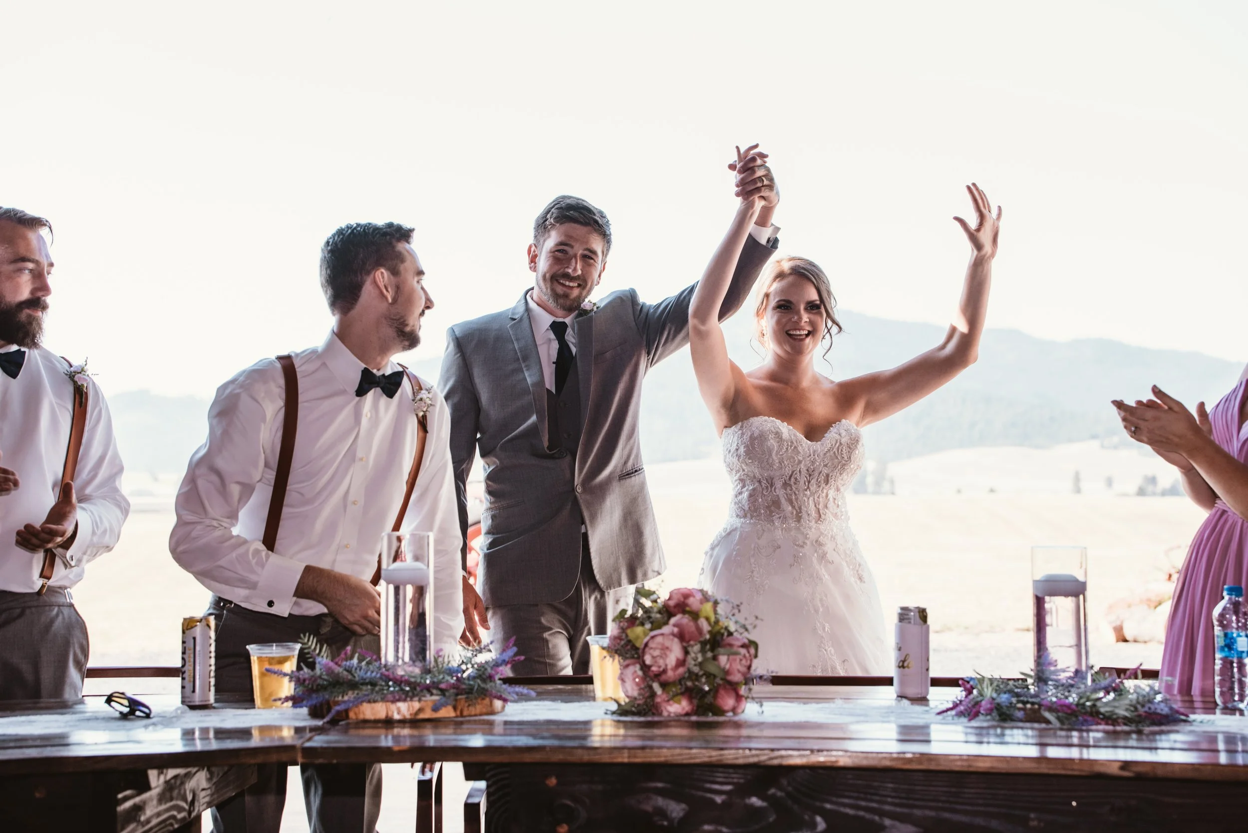 Bride and groom celebrating at a wedding reception with groomsmen and bridesmaid, holding hands up.