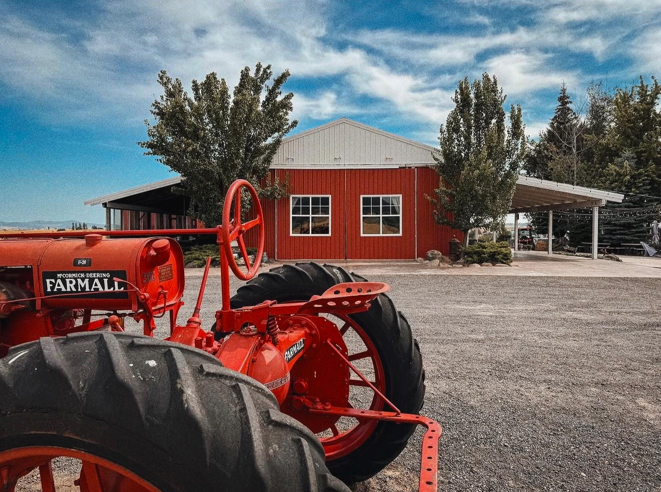 Red vintage tractor with large black tires parked in front of a red barn with white trim, surrounded by trees and under a partly cloudy sky.