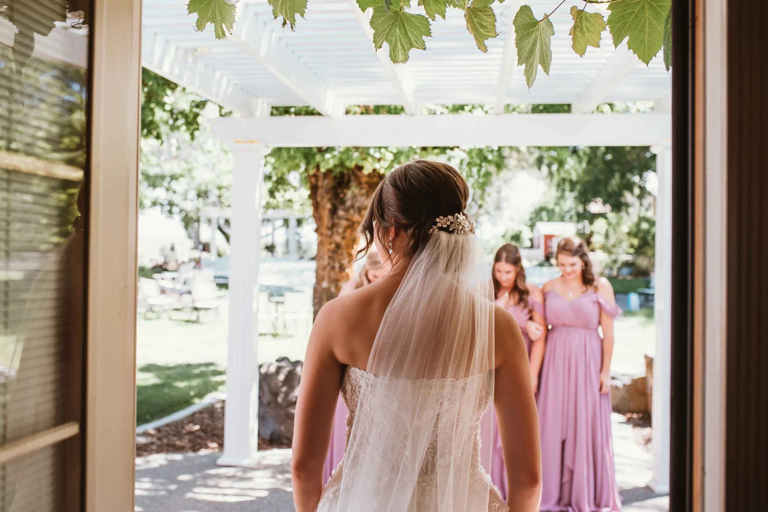 Bride standing in front of bridesmaids outside under a pergola with green leaves.