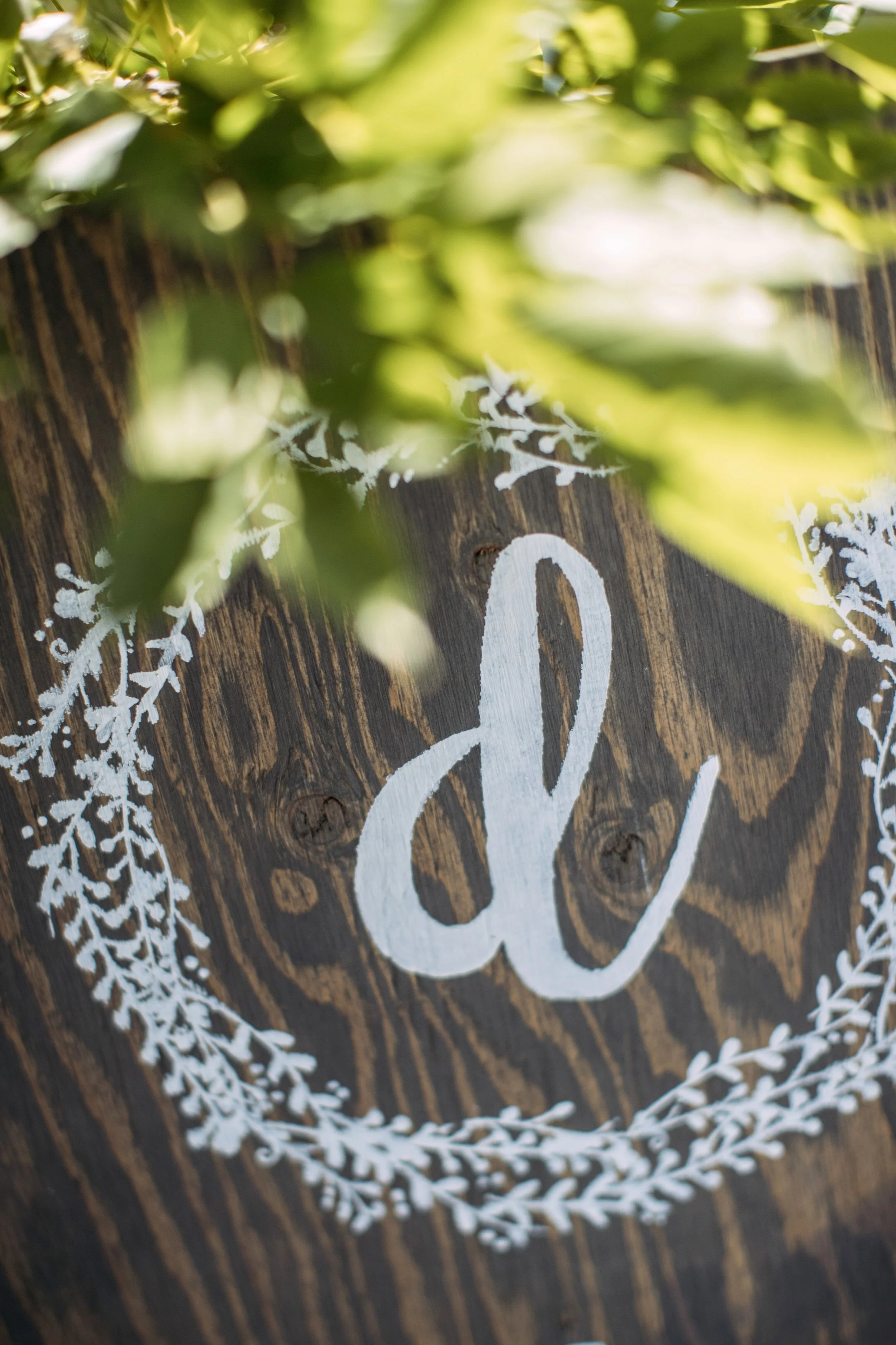 Decorative wooden sign with a letter 'd' and white floral wreath, partially covered by green leaves.