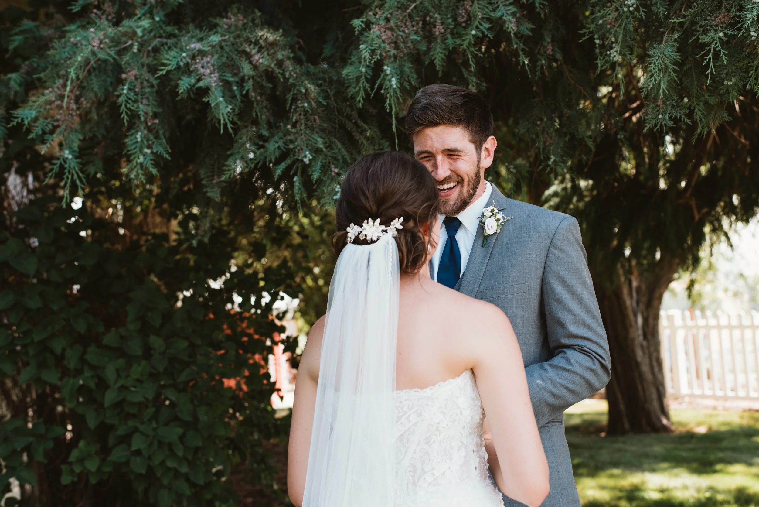 Bride in white dress facing groom in gray suit under tree, outdoors.