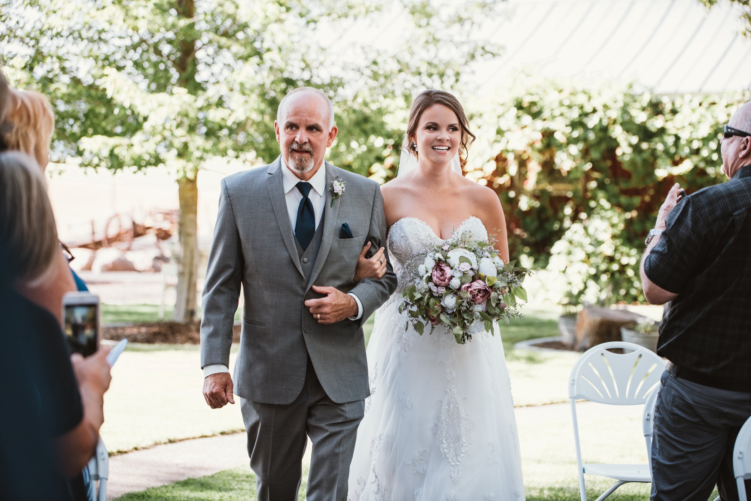 Bride walking down the aisle with her father, both smiling, during an outdoor wedding ceremony. The bride is holding a bouquet, and guests are watching and taking photos.