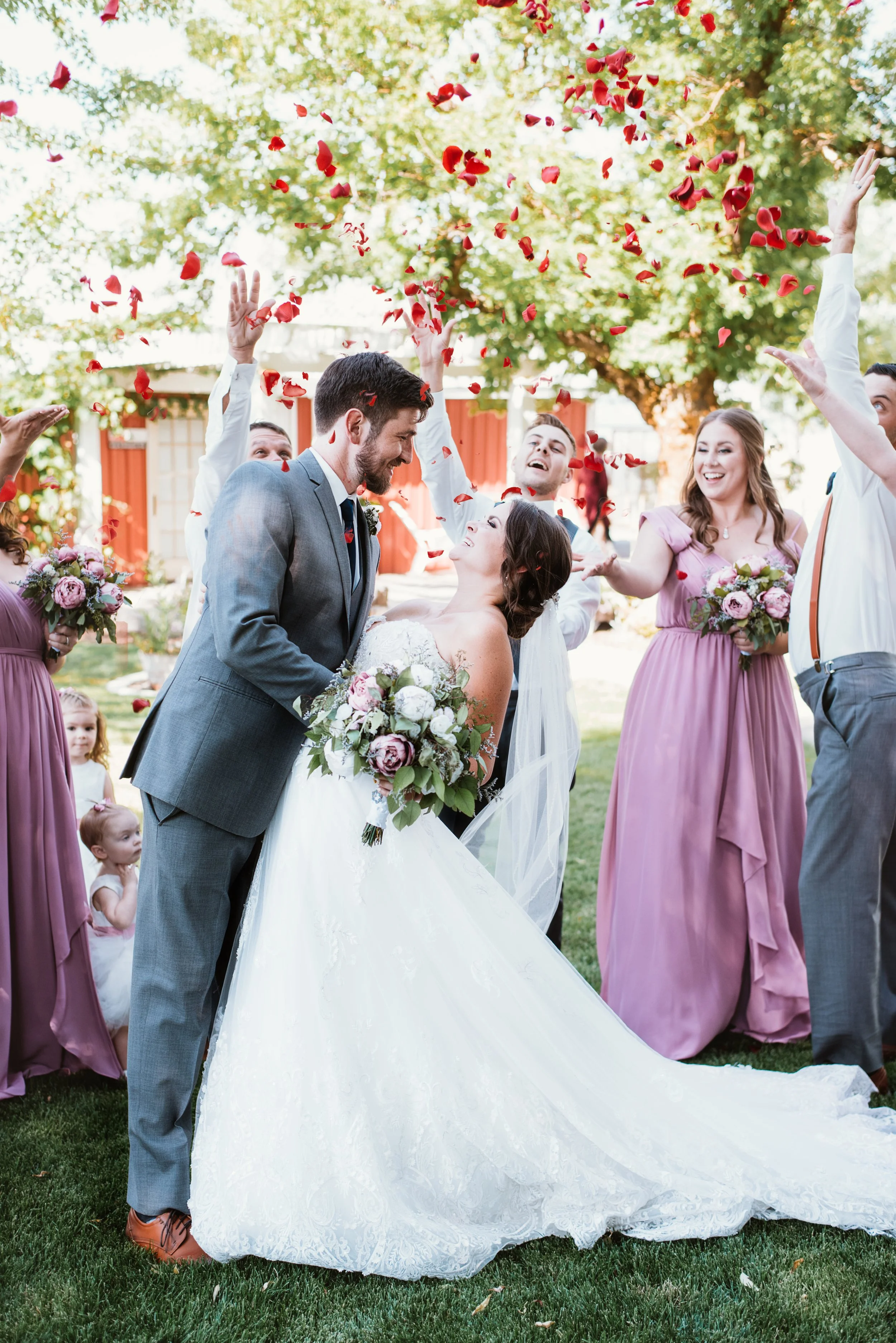Bride and groom at outdoor wedding surrounded by flower petals and bridal party.