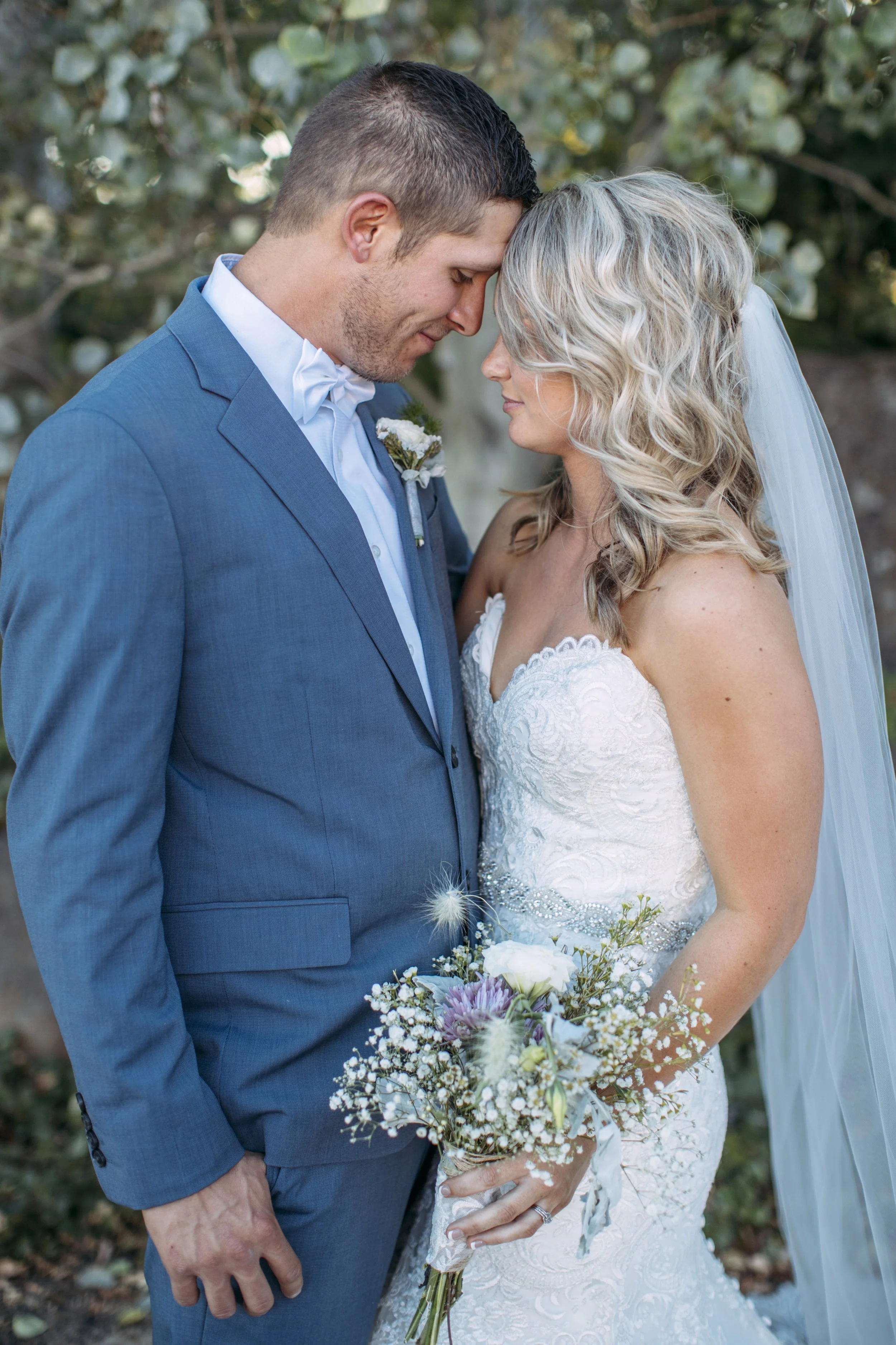 Bride and groom touching foreheads, wearing wedding attire, with a bouquet, in an outdoor setting.