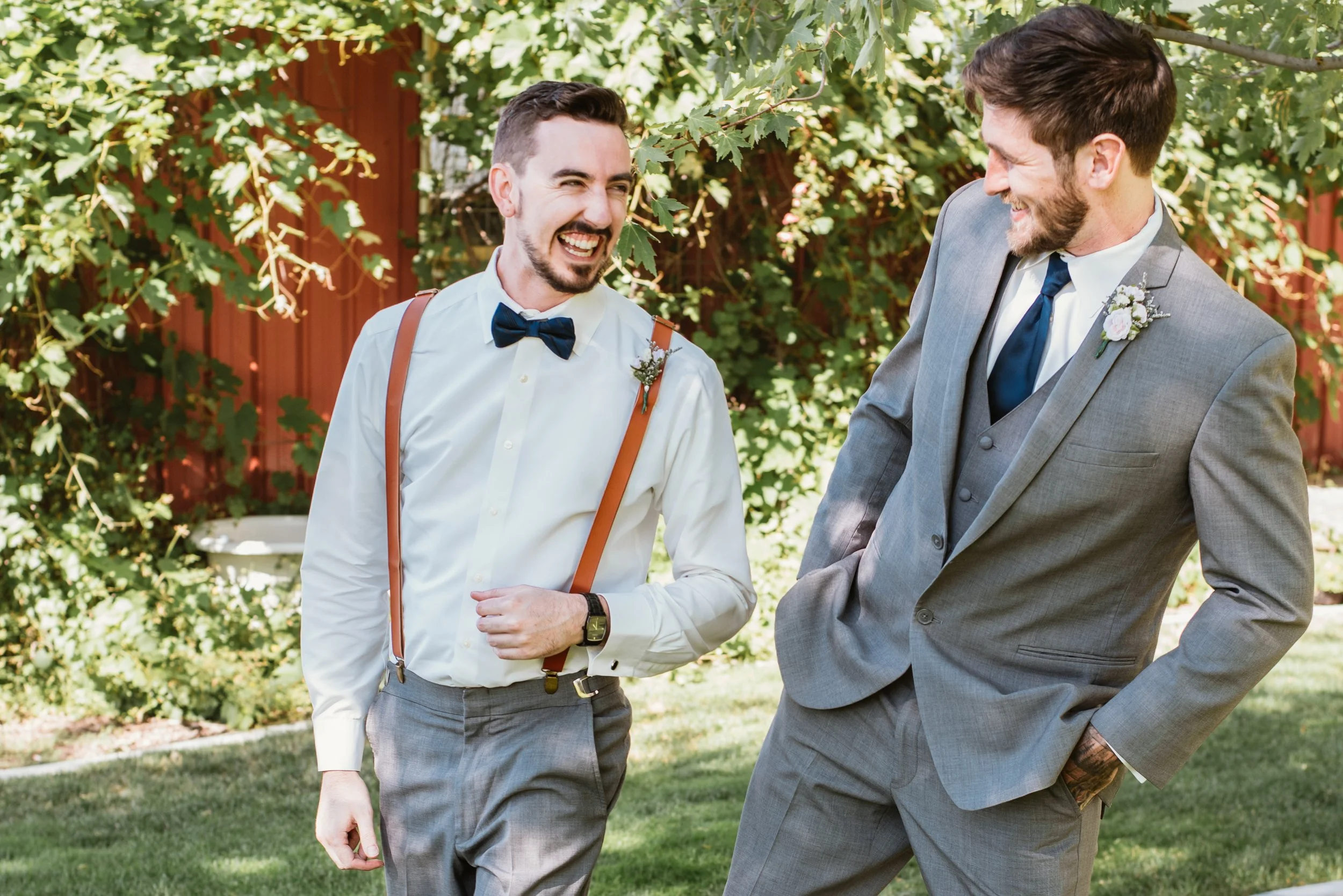 Two men smiling and laughing outdoors, dressed in formal attire. One wears a white shirt with suspenders and a bow tie, while the other is in a gray suit and tie. They are surrounded by greenery and sunlight.