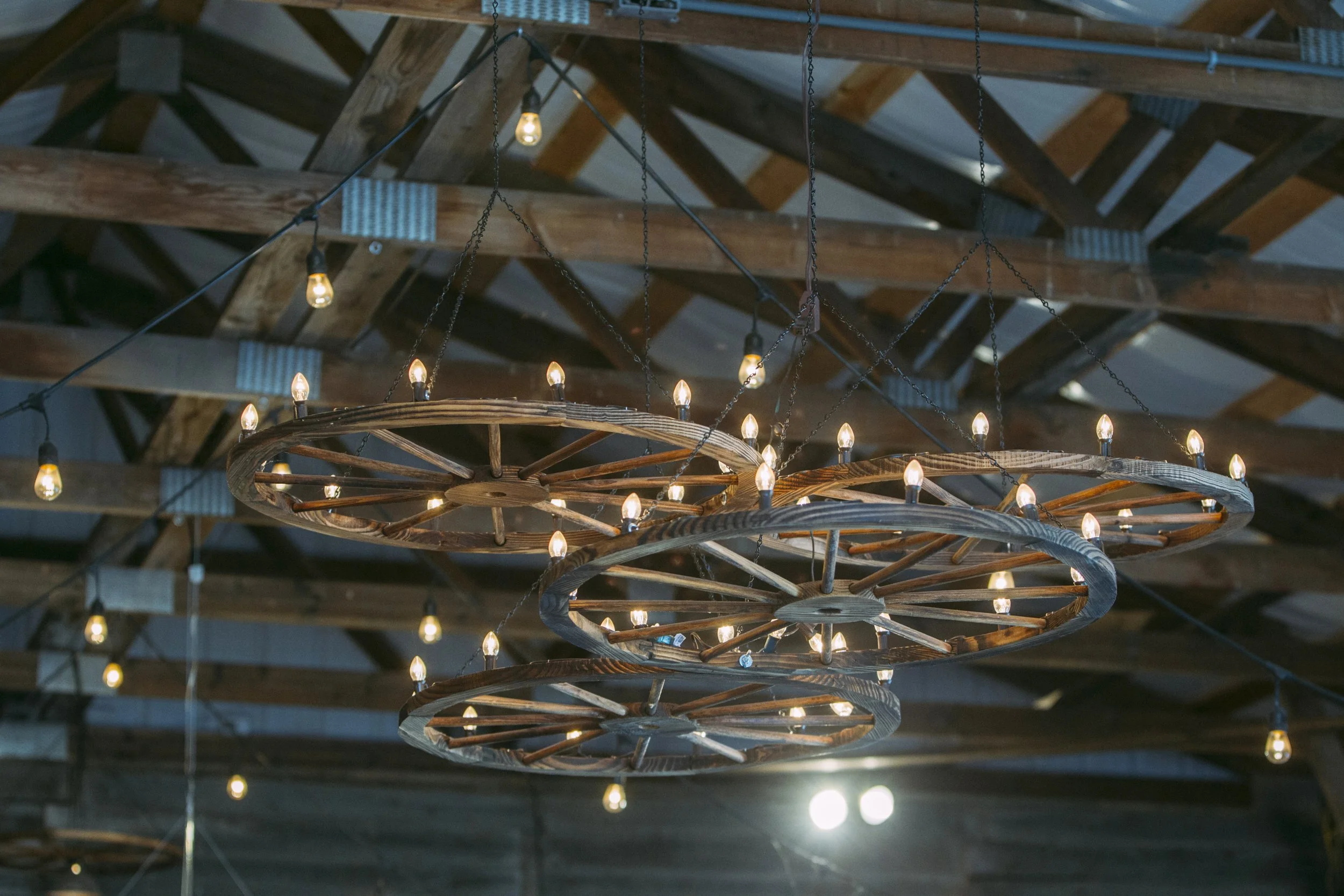 Rustic chandelier made from wagon wheels and Edison bulbs, hanging from a wooden barn ceiling.