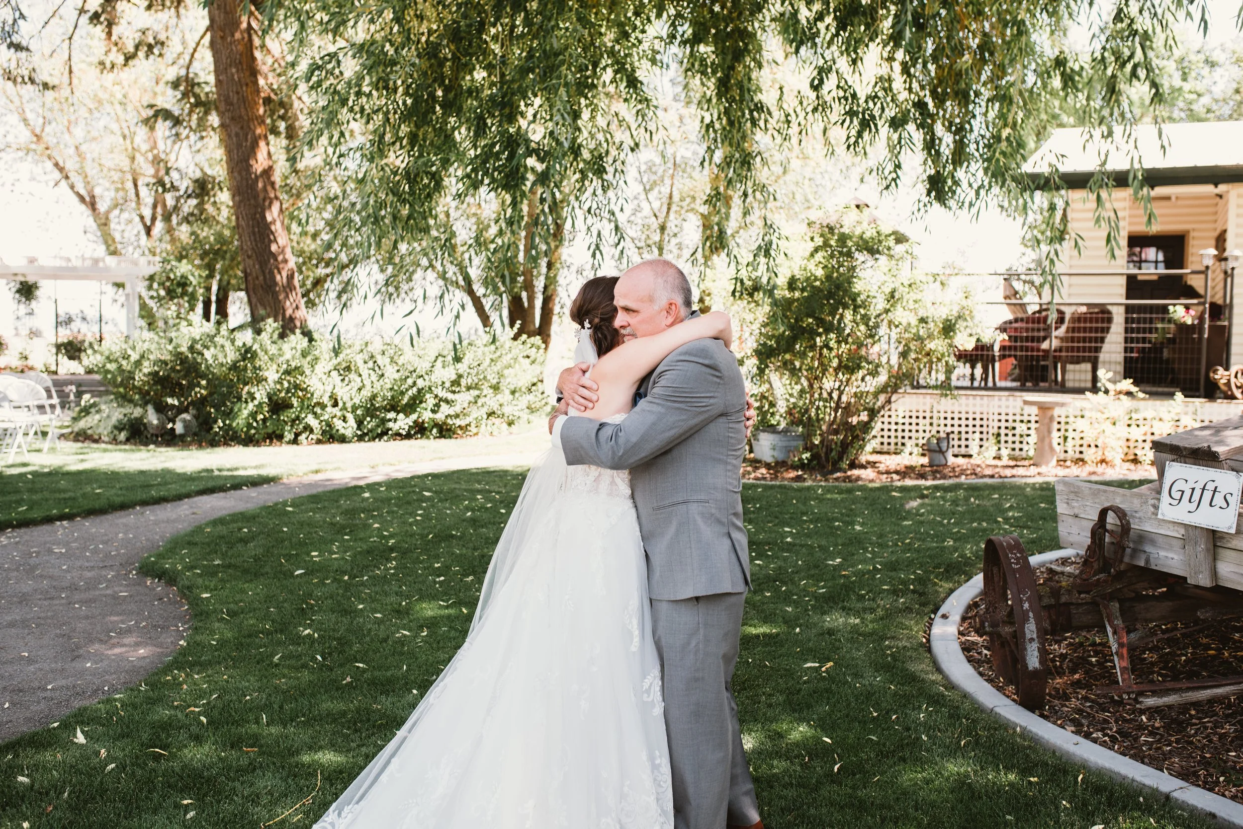 A bride in a white wedding dress hugs an elderly man in a gray suit outdoors, surrounded by trees and greenery. A wooden cart with a 'Gifts' sign is visible on the right.