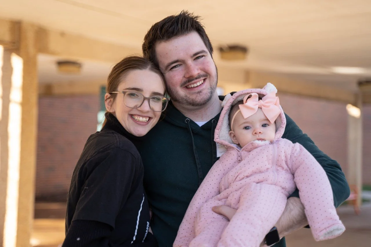 A smiling couple is holding a baby dressed in a pink outfit with a bow. They are standing outdoors.
