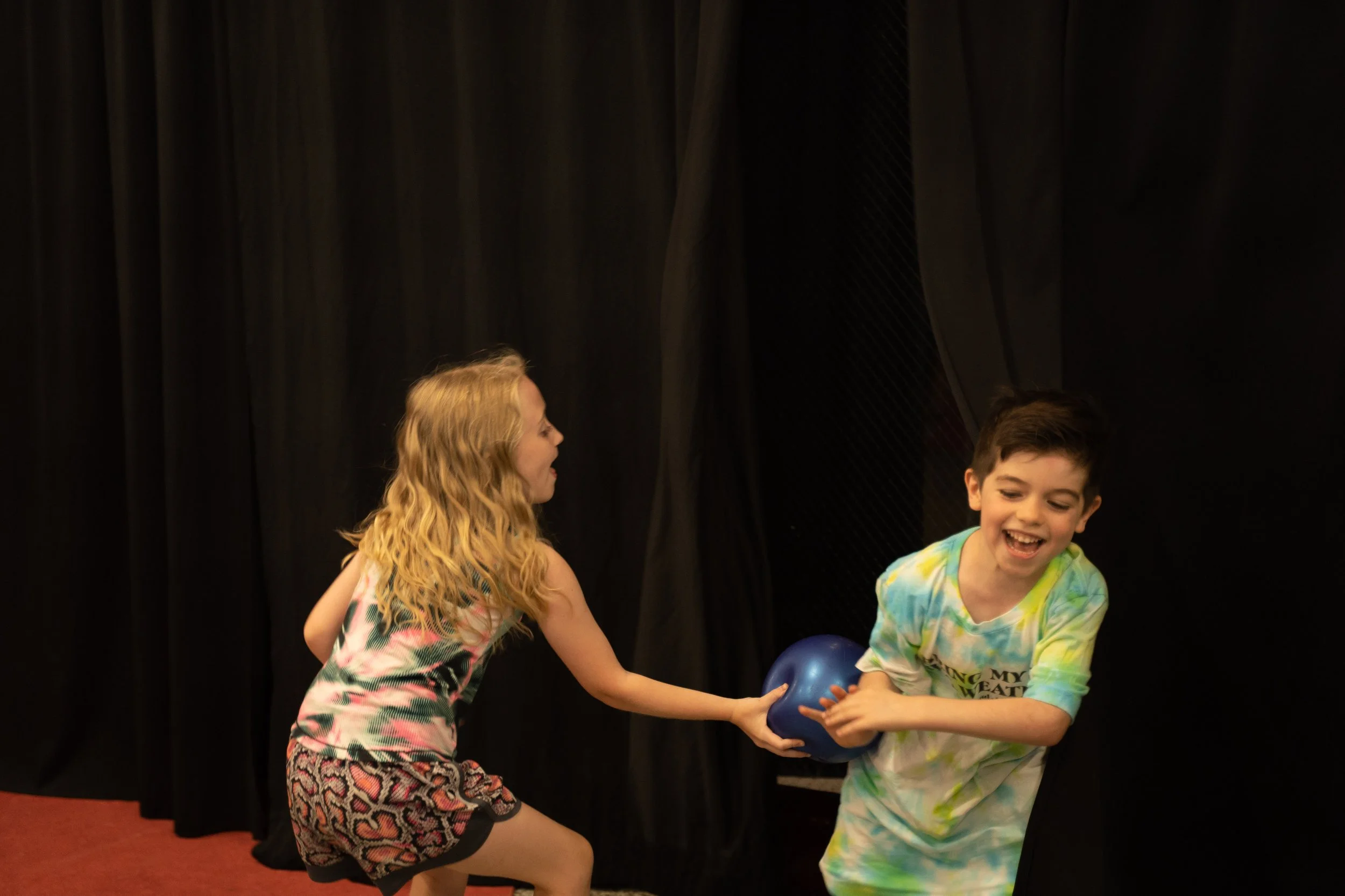 Two children playing with a blue ball indoors against a black curtain background. The girl with long blonde hair is reaching for the ball from the boy, who is wearing a tie-dye shirt and smiling.