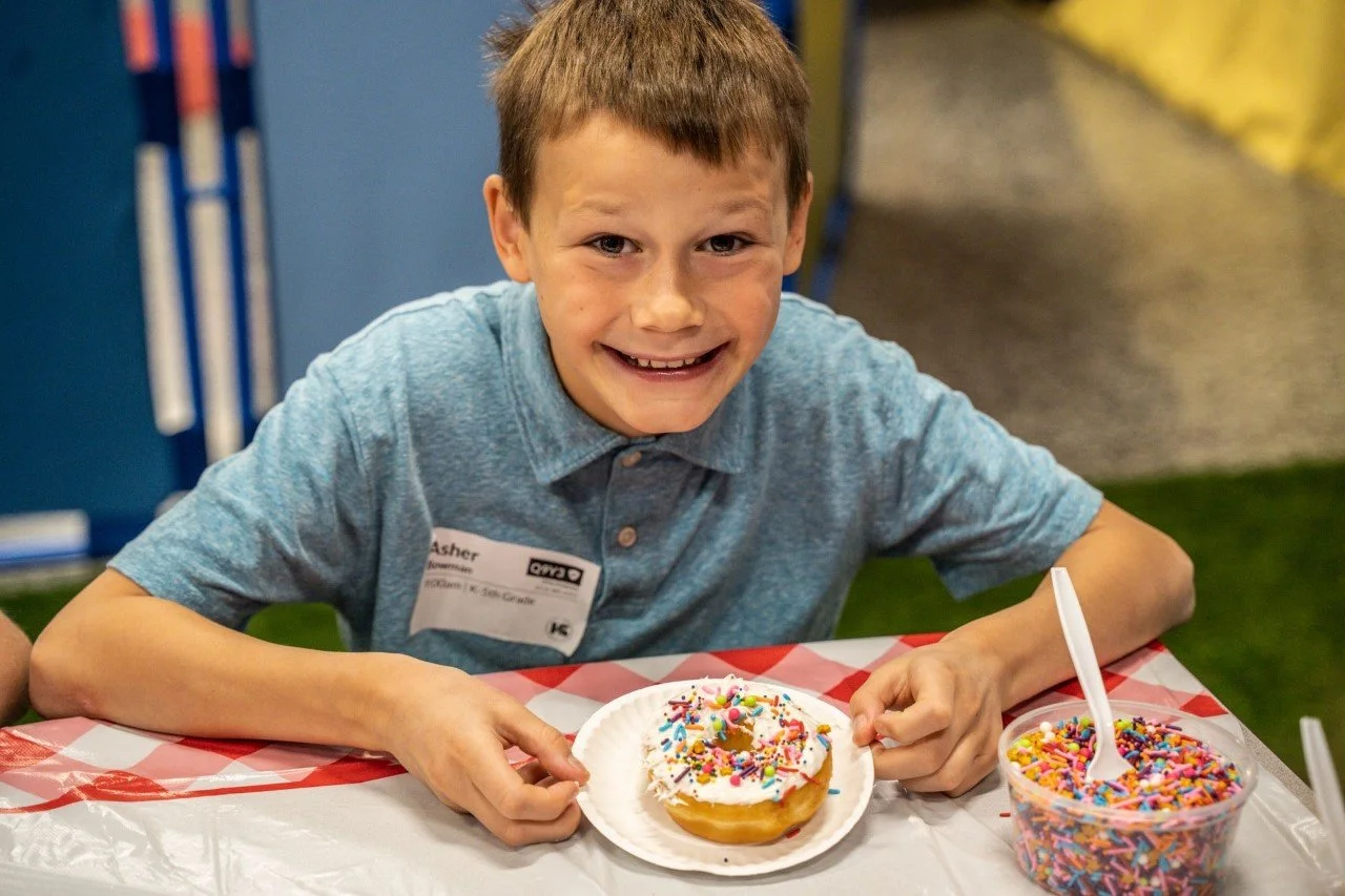 Boy smiling at a table with a doughnut covered in white frosting and colorful sprinkles, next to a container of sprinkles.
