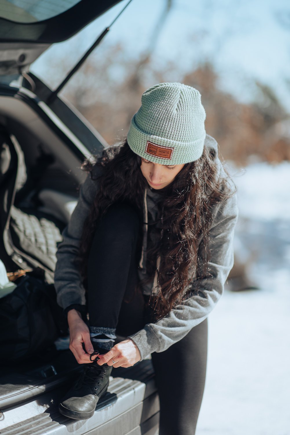 Professional lifestyle photography of young model with long brown hair wearing a teal Akinz Boardwear beanie and gray Colorado sweatshirt in the snowy mountains in Colorado Springs, Colorado