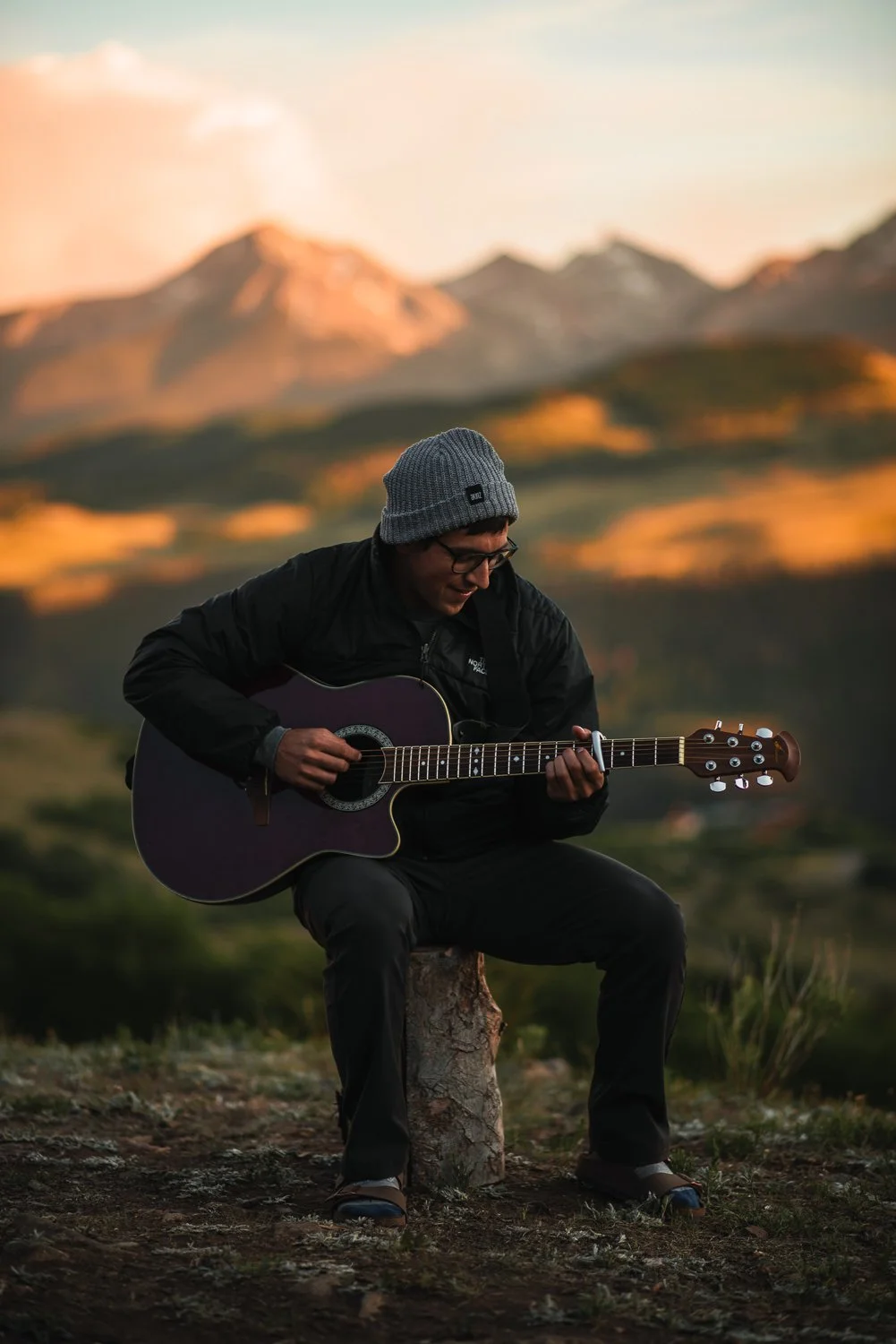 Outdoor product photography of a man sitting on a log playing guitar, wearing Akinz Boardwear gear while camping in the mountains in Telluride, CO.