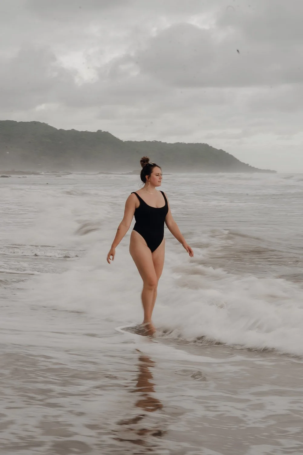 Travel image of a girl wearing a black one piece swimsuit walking into the ocean on an overcast sunset in Santa Teresa, Costa Rica