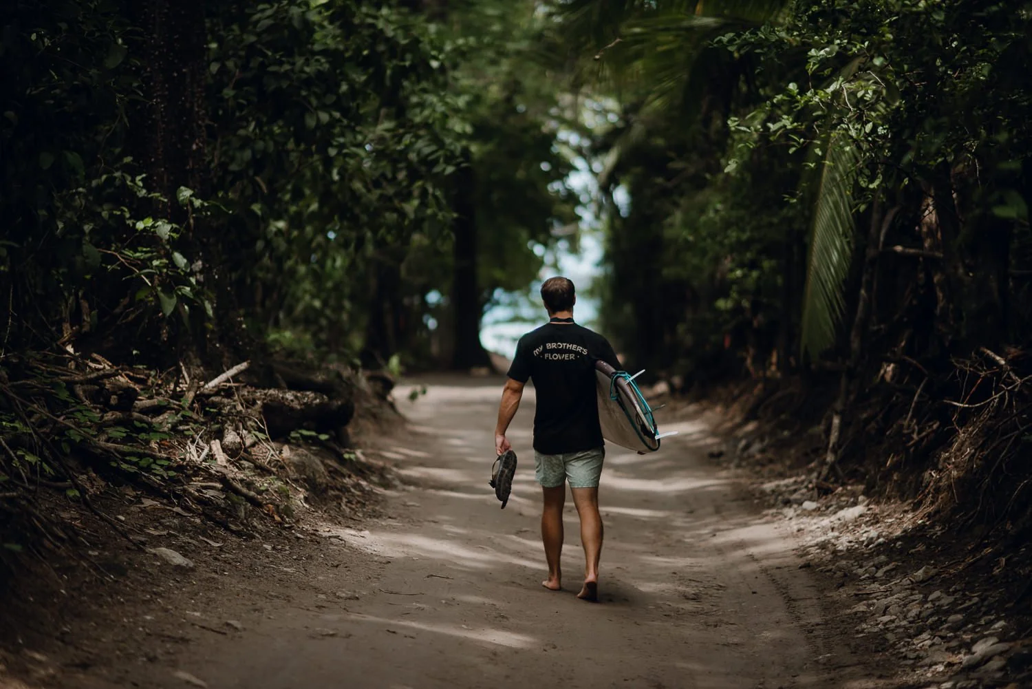 Adventure photography of a young man holding a surfboard walking along lush rainforest path toward the beach