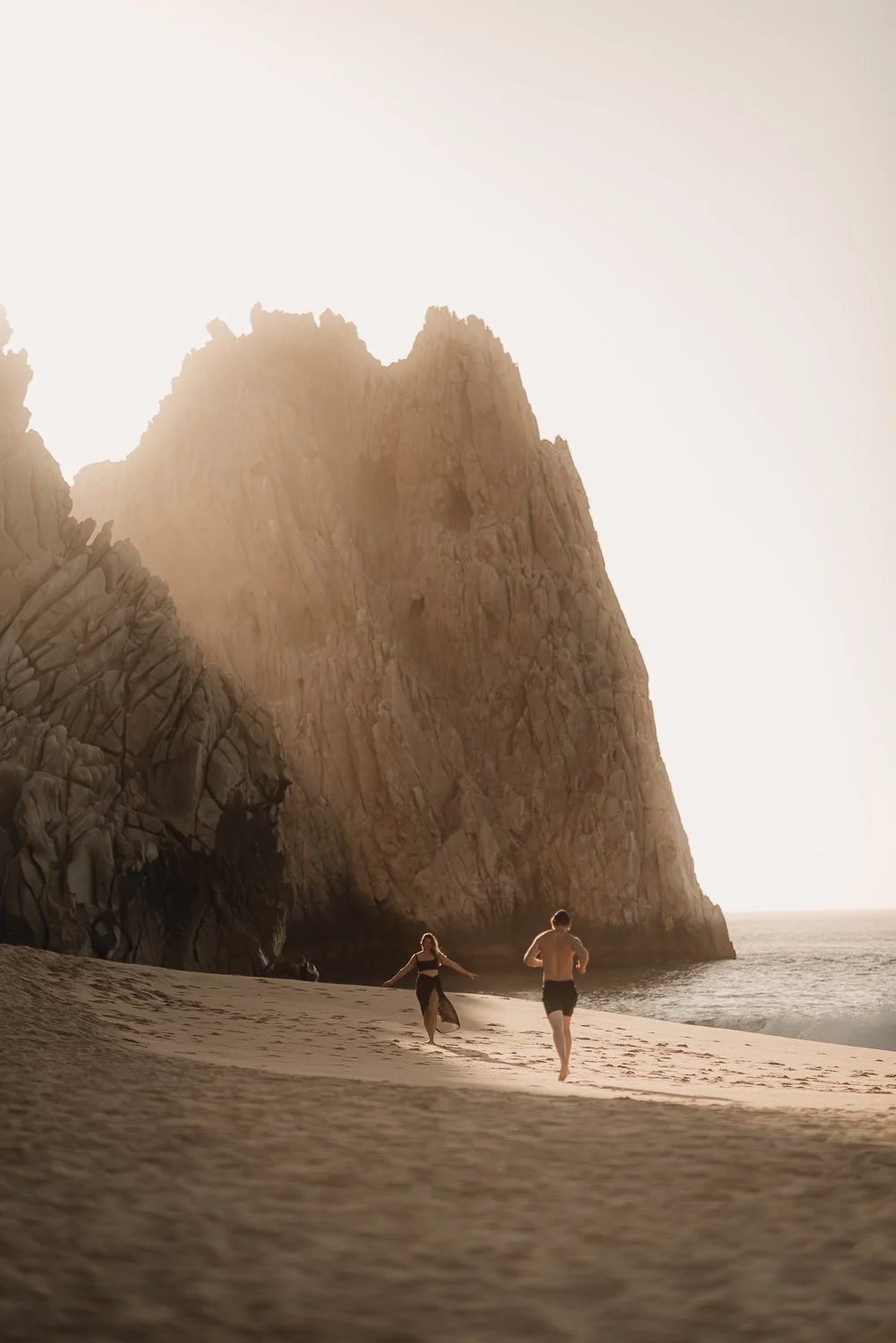 Travel photography of a couple walking on the shore in front of unique rock formations during hazy sunrise at Lover's Beach in Cabo San Lucas, Mexico
