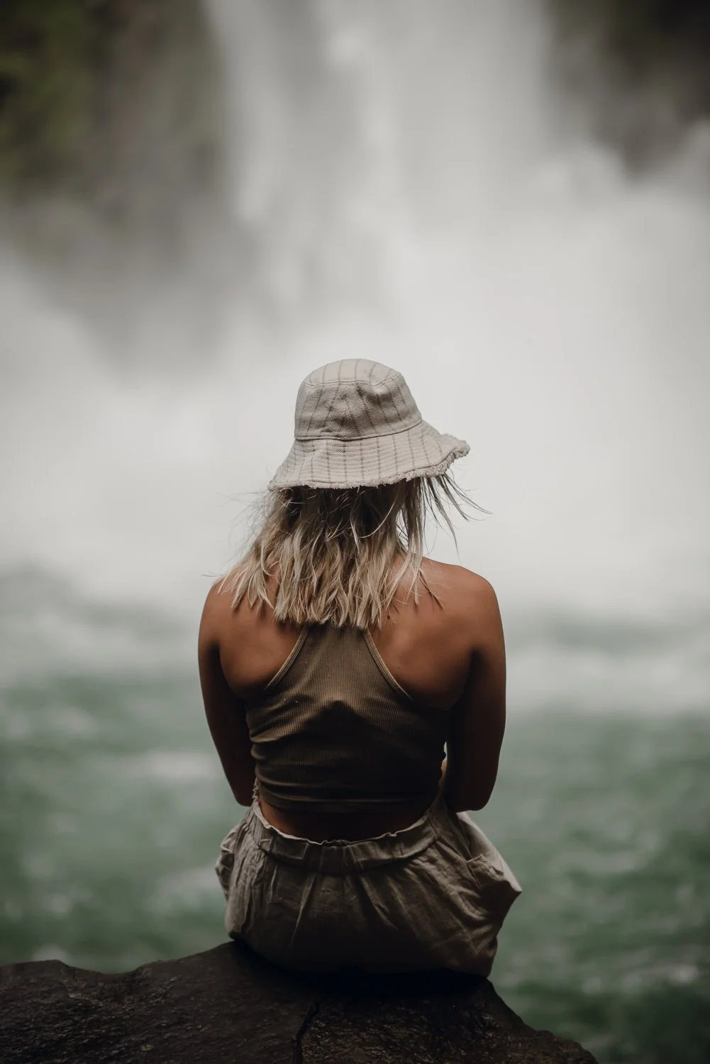 Lifestyle portrait of a blonde girl wearing a bucket hat sitting in front of La Fortuna waterfall in Costa Rica