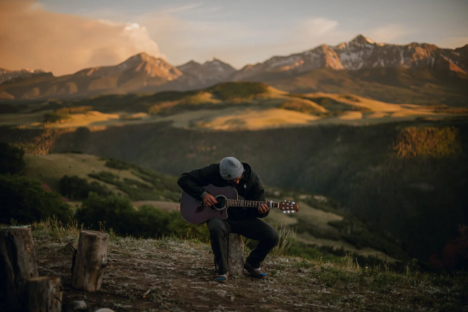 Outdoor product photography of a man sitting on a log playing guitar, wearing Akinz Boardwear gear while camping in the mountains in Telluride, CO.