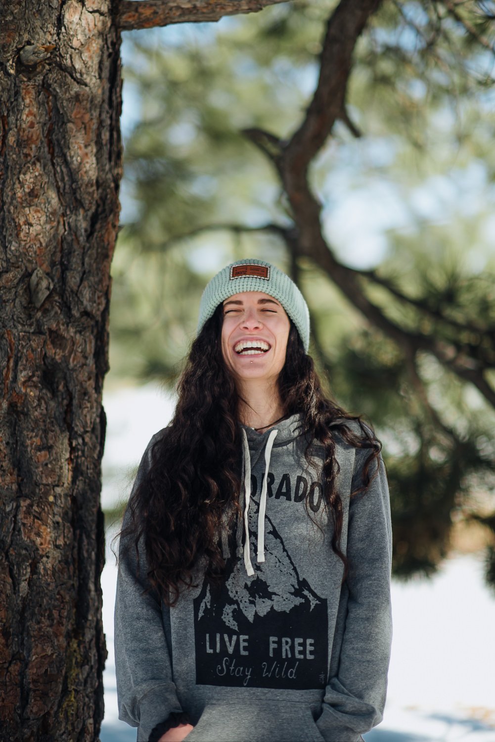 Professional lifestyle photography of young model with long brown hair wearing a teal Akinz Boardwear beanie and gray Colorado sweatshirt in the snowy mountains in Colorado Springs, Colorado