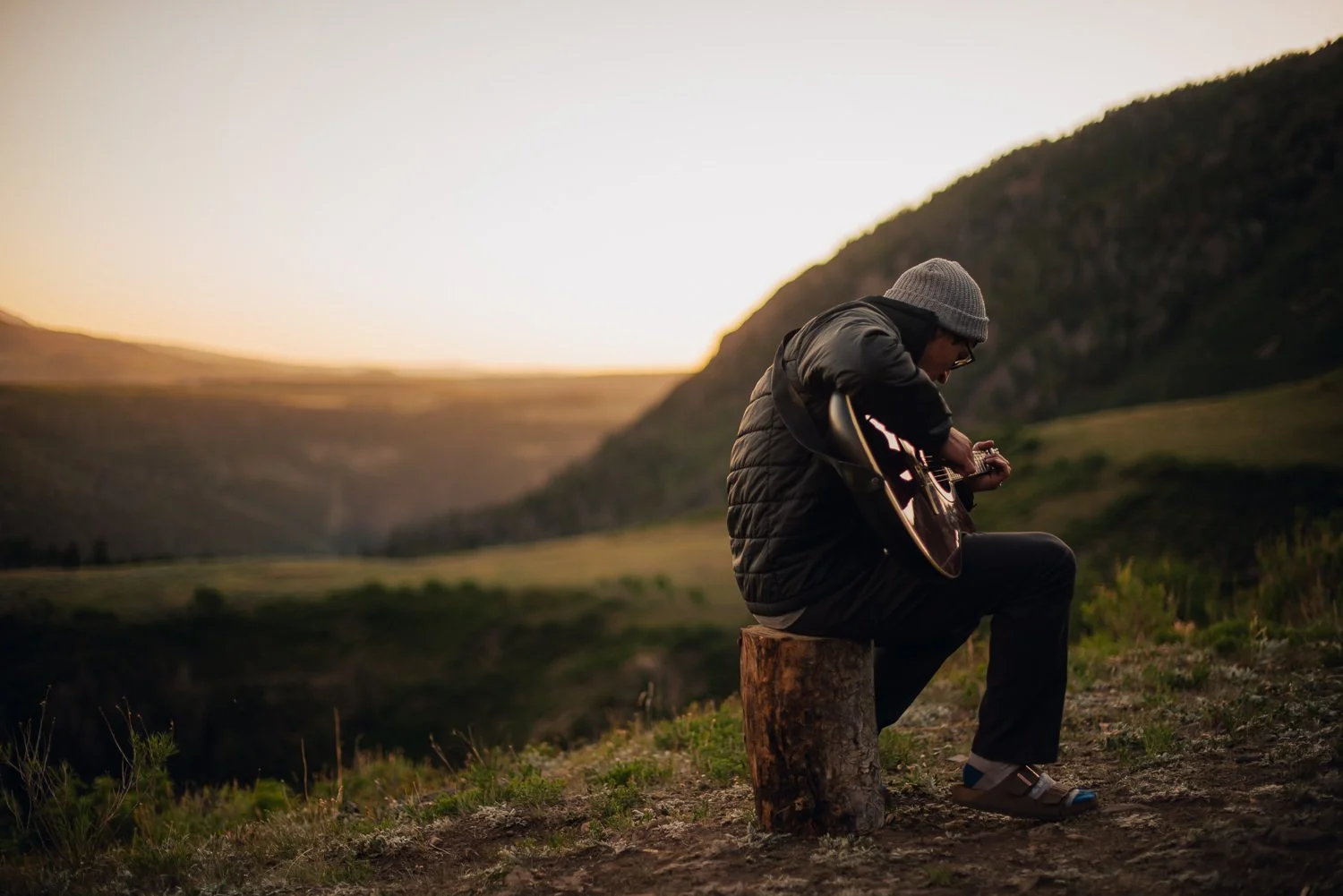 Outdoor product photography of a man sitting on a log playing guitar, wearing Akinz Boardwear gear while camping in the mountains in Telluride, CO.