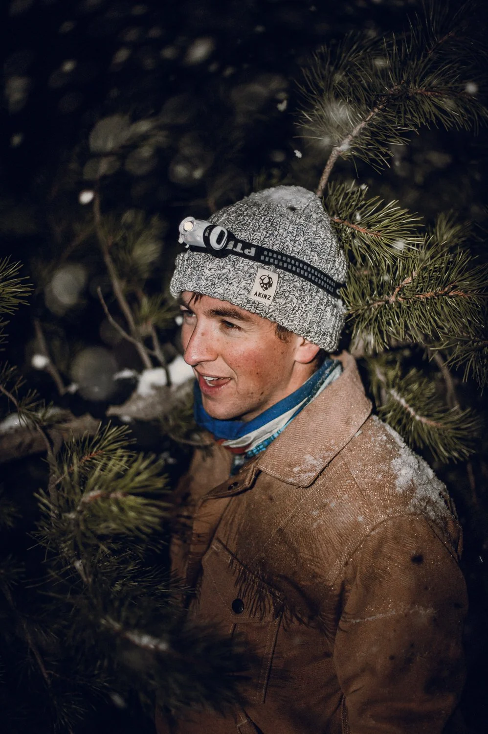 Lifestyle brand photography of young man wearing an Akinz beanie in the woods at night, while carrying a cut-down christmas tree through the snow