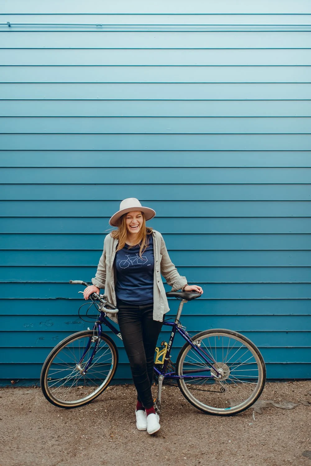 Lifestyle photography of a cute blonde girl in a stylish hat leaning against a bike in front of a blue wall in Colorado Springs, CO for Akinz Boardwear