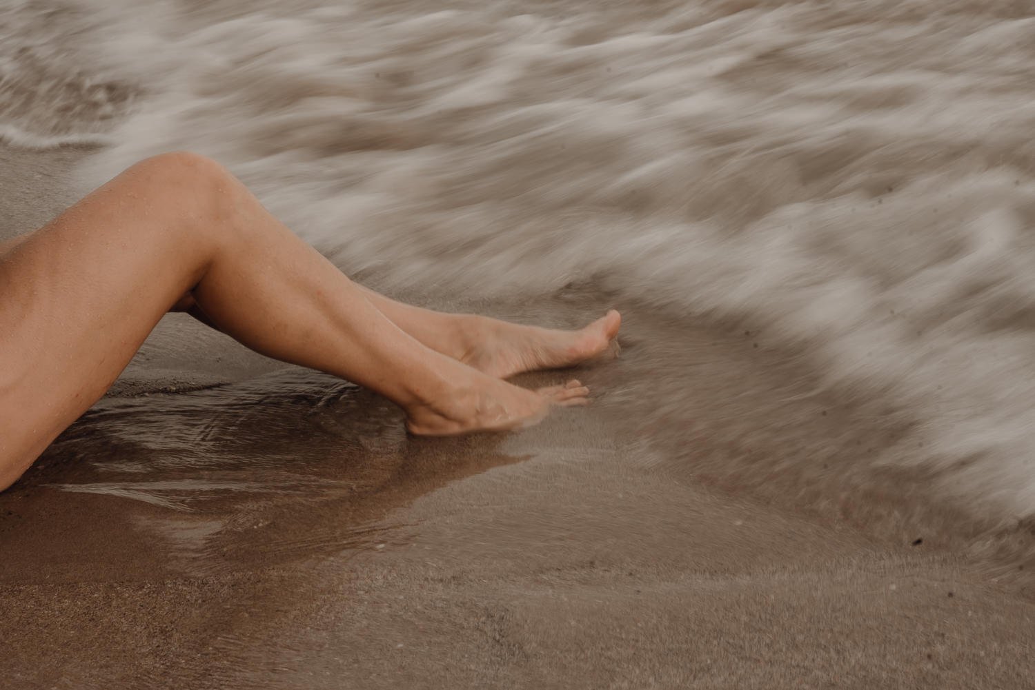 Slow shutter speed travel photo of feet in sand as waves wash up the shore on Santa Teresa Beach in Costa Rica