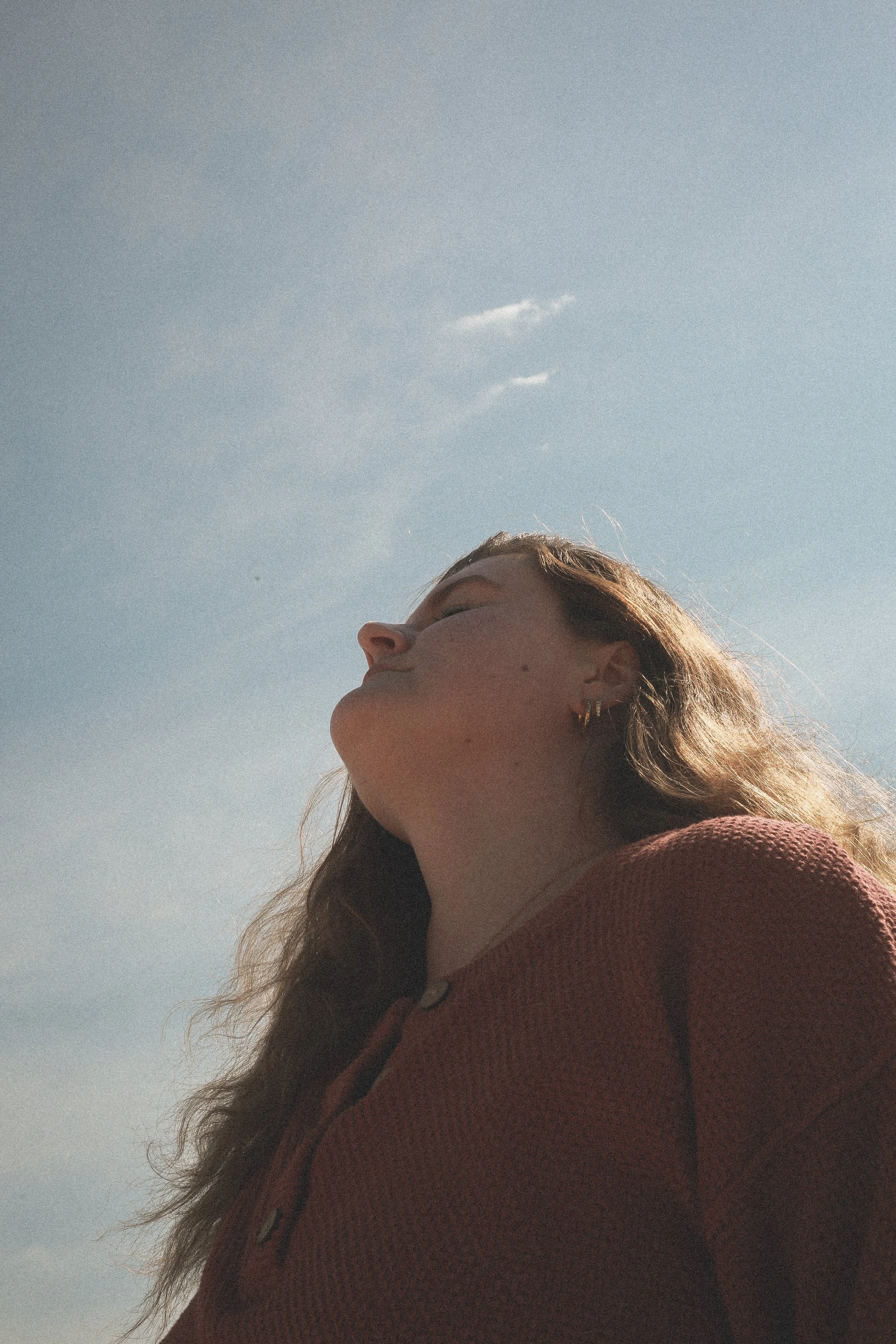 A woman with closed eyes and a slight smile standing outdoors on a sunny day, wearing a reddish-brown top, with her head tilted back and hair flowing.