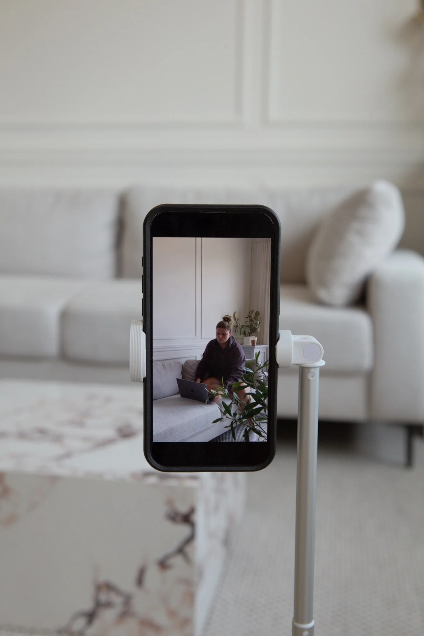 A smartphone mounted on a tripod showing a woman sitting on a sofa using a laptop in a bright living room with white walls, a plant, and a marble table in the foreground.