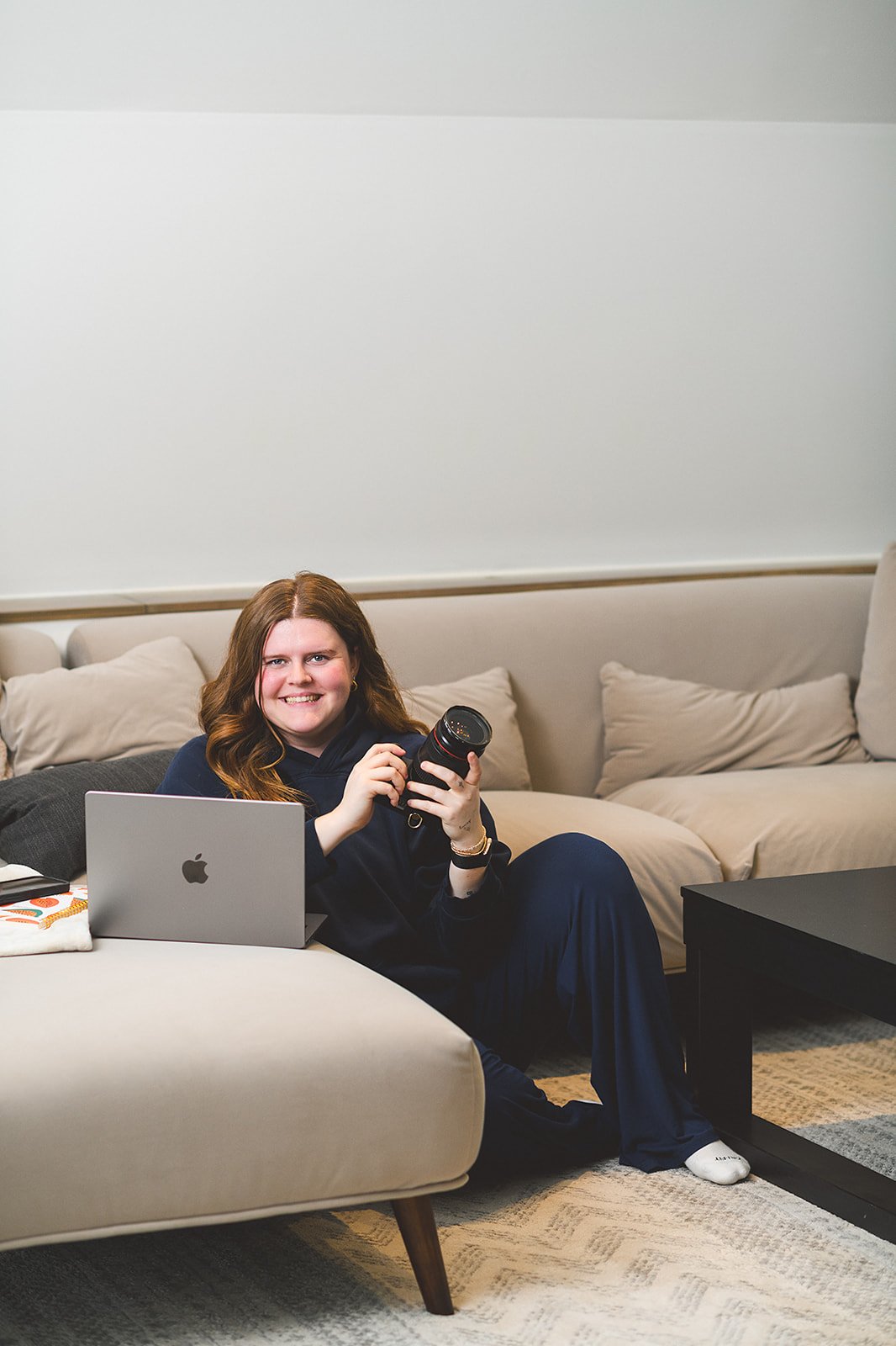 A woman with long red hair sitting on a beige sofa, holding a camera in one hand and a laptop on her lap, smiling at the camera.