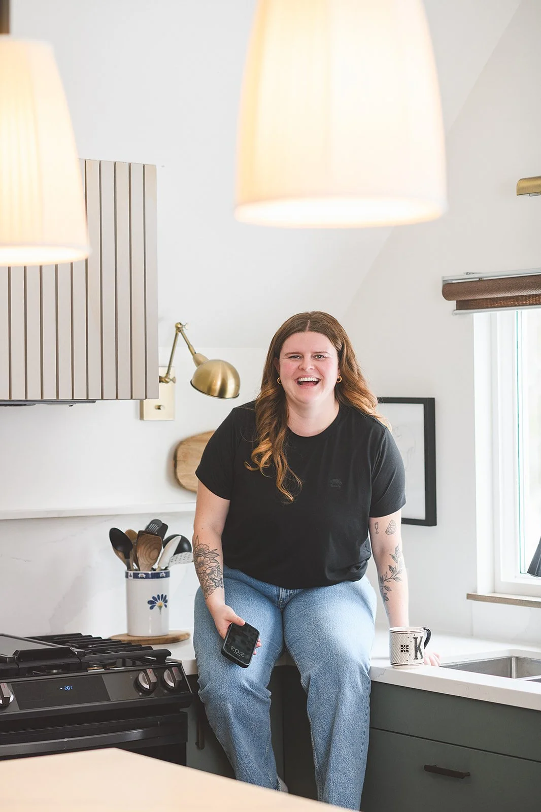 A woman with long wavy brown hair, tattoos on her arms, wearing a black t-shirt and blue jeans, sitting on a kitchen counter, smiling and holding a smartphone. The kitchen features pendant lights, a window, and various utensils.