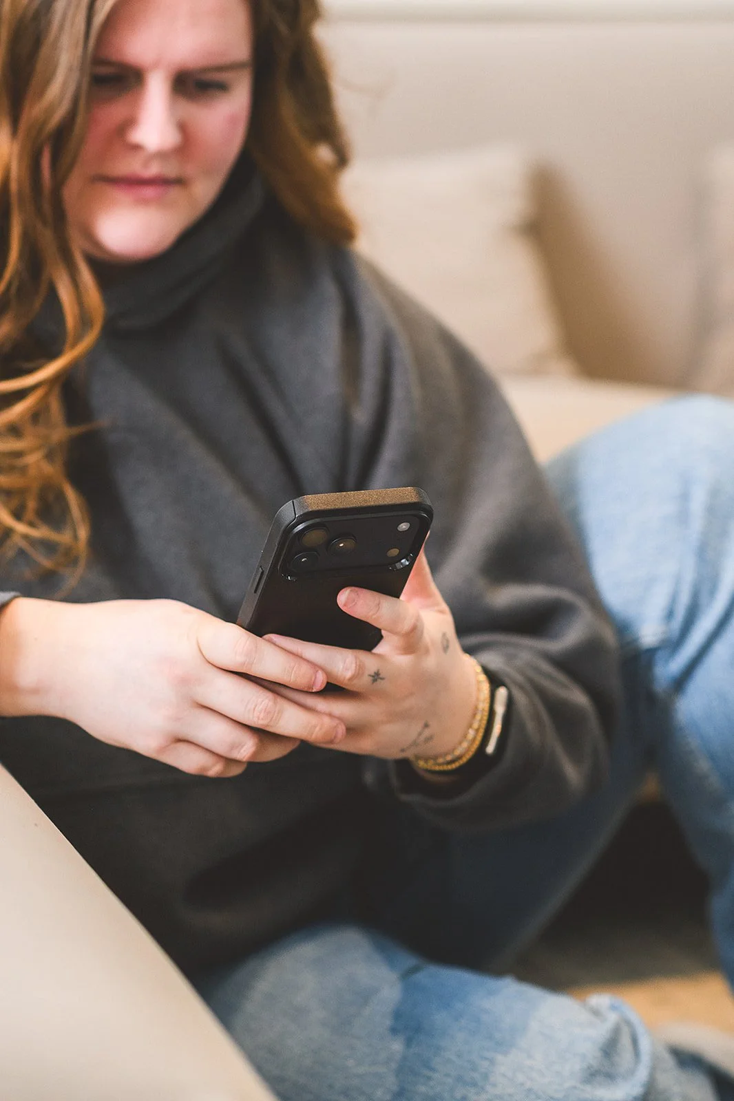 A woman with wavy red hair wearing a dark gray sweater and jeans is sitting on a couch, looking at her smartphone.