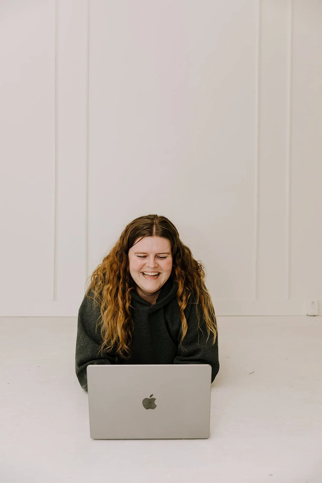 A young woman with long, curly hair smiling while looking at her laptop, which is placed on the floor in front of her.