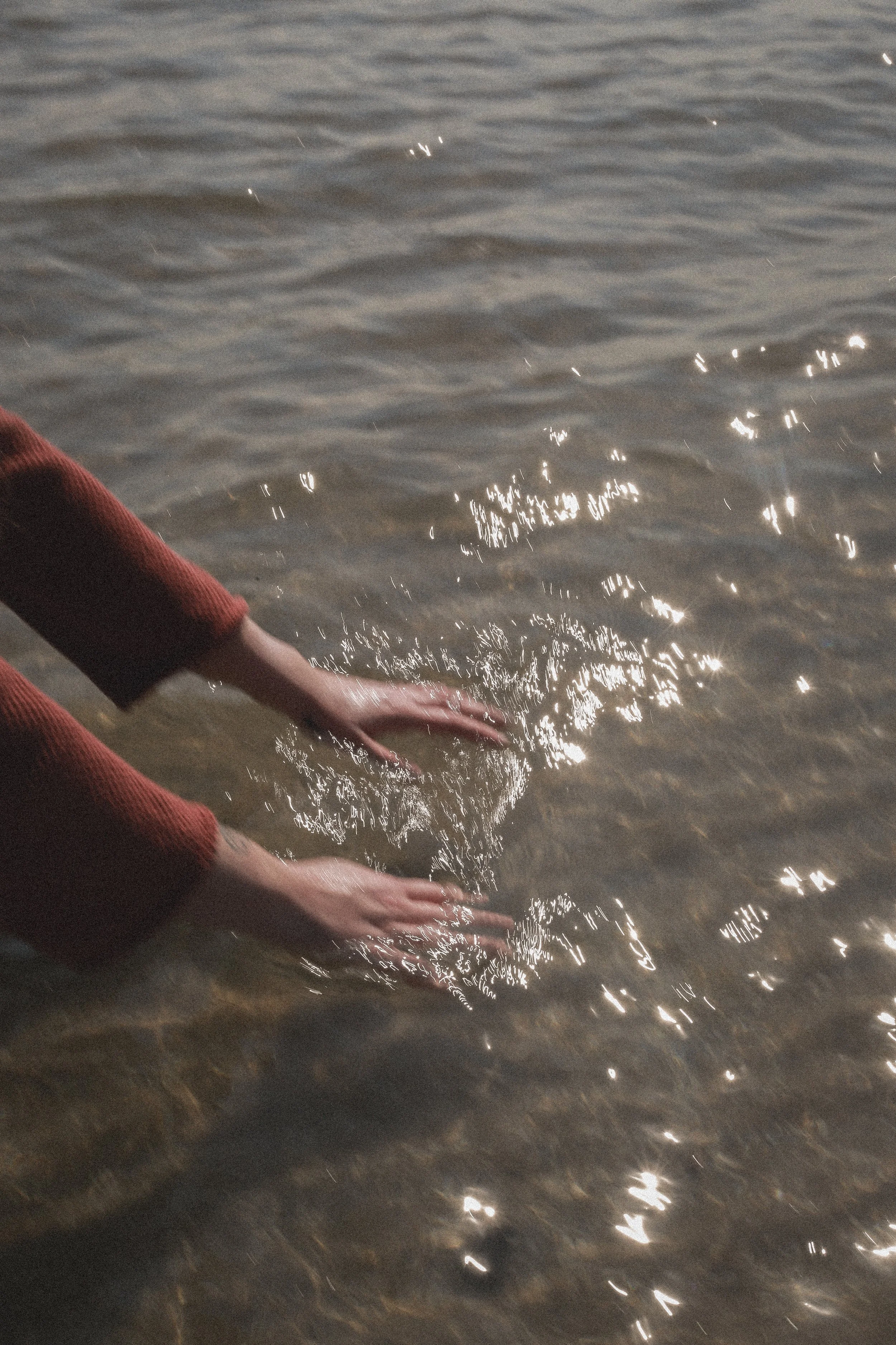 Person with long sleeves touching the water near the shoreline at sunset, with light reflecting on the surface.