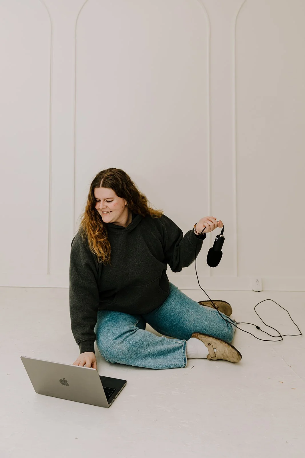 A woman with long brown hair wearing a dark gray hoodie, blue jeans, and beige boots, sitting on the floor in a white room. She is holding a microphone and using a laptop, smiling and looking at the laptop.
