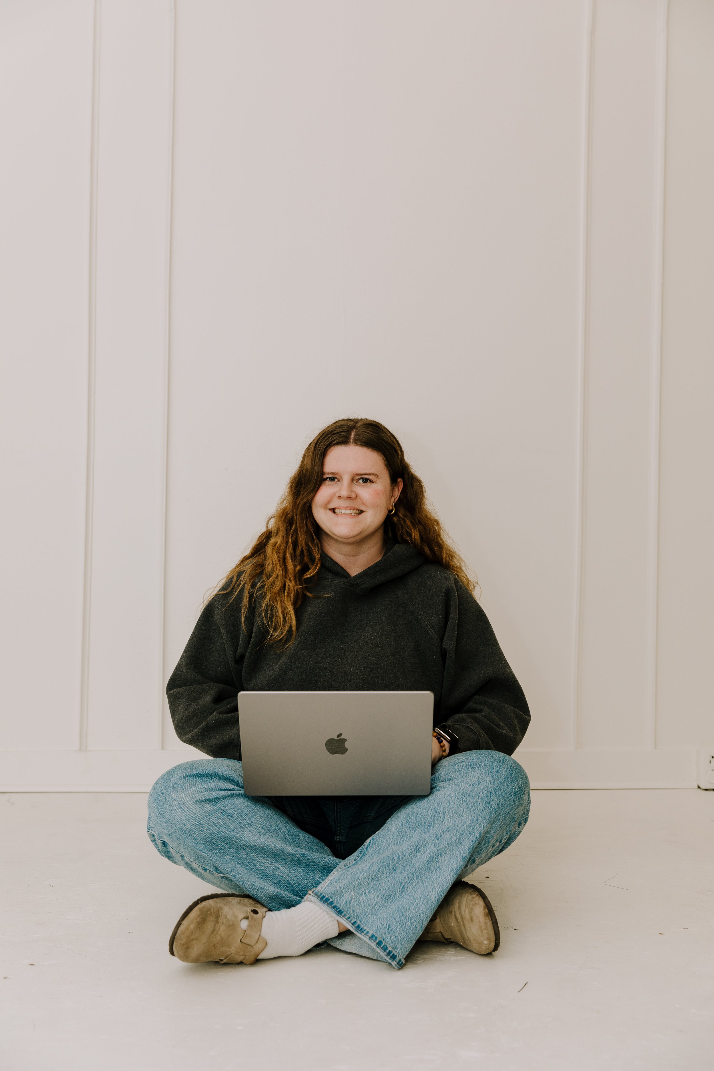 A young woman with long curly hair sitting cross-legged on the floor with a laptop on her lap against a white wall.