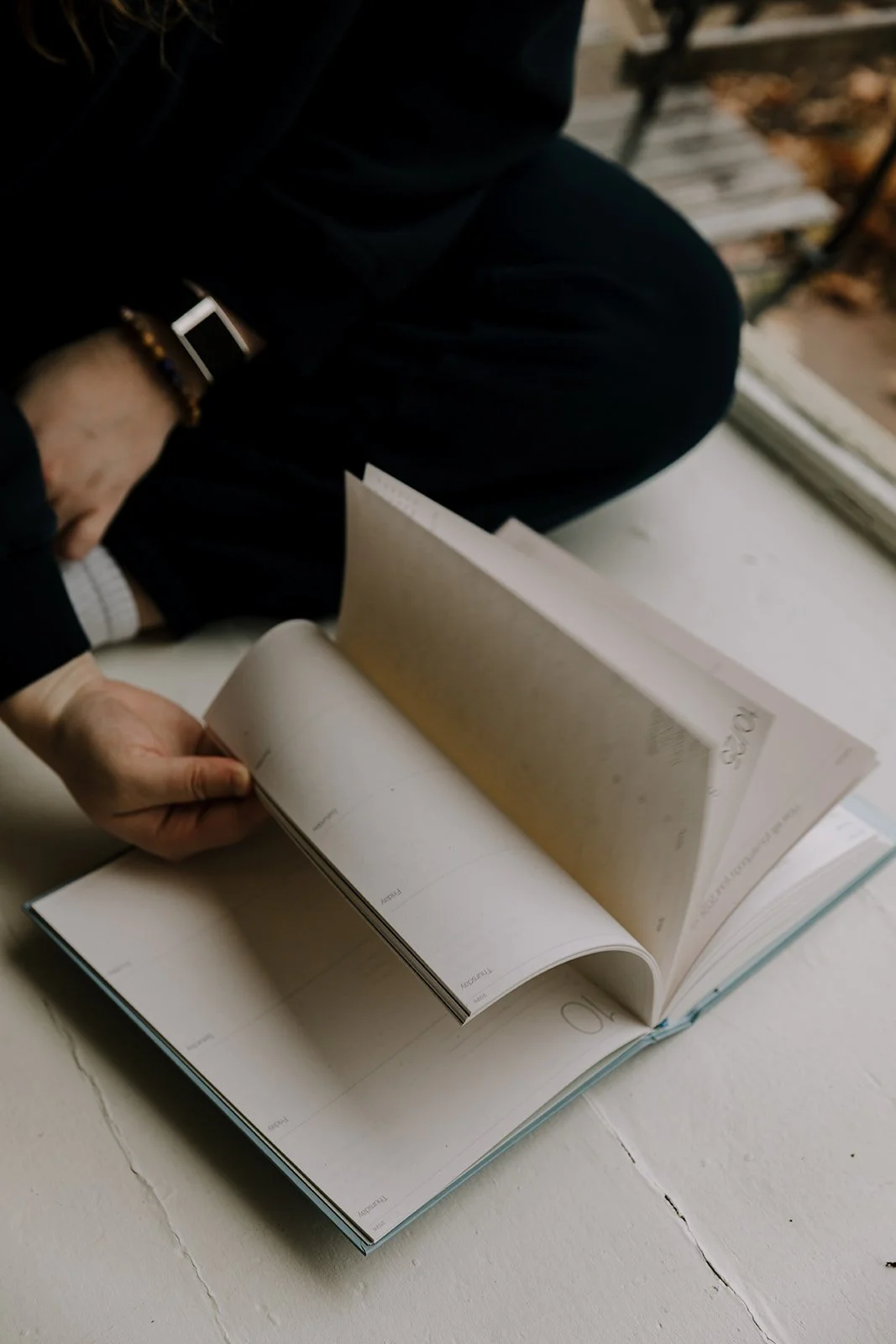 Person flipping pages of a planner or journal on a white table, near a window.