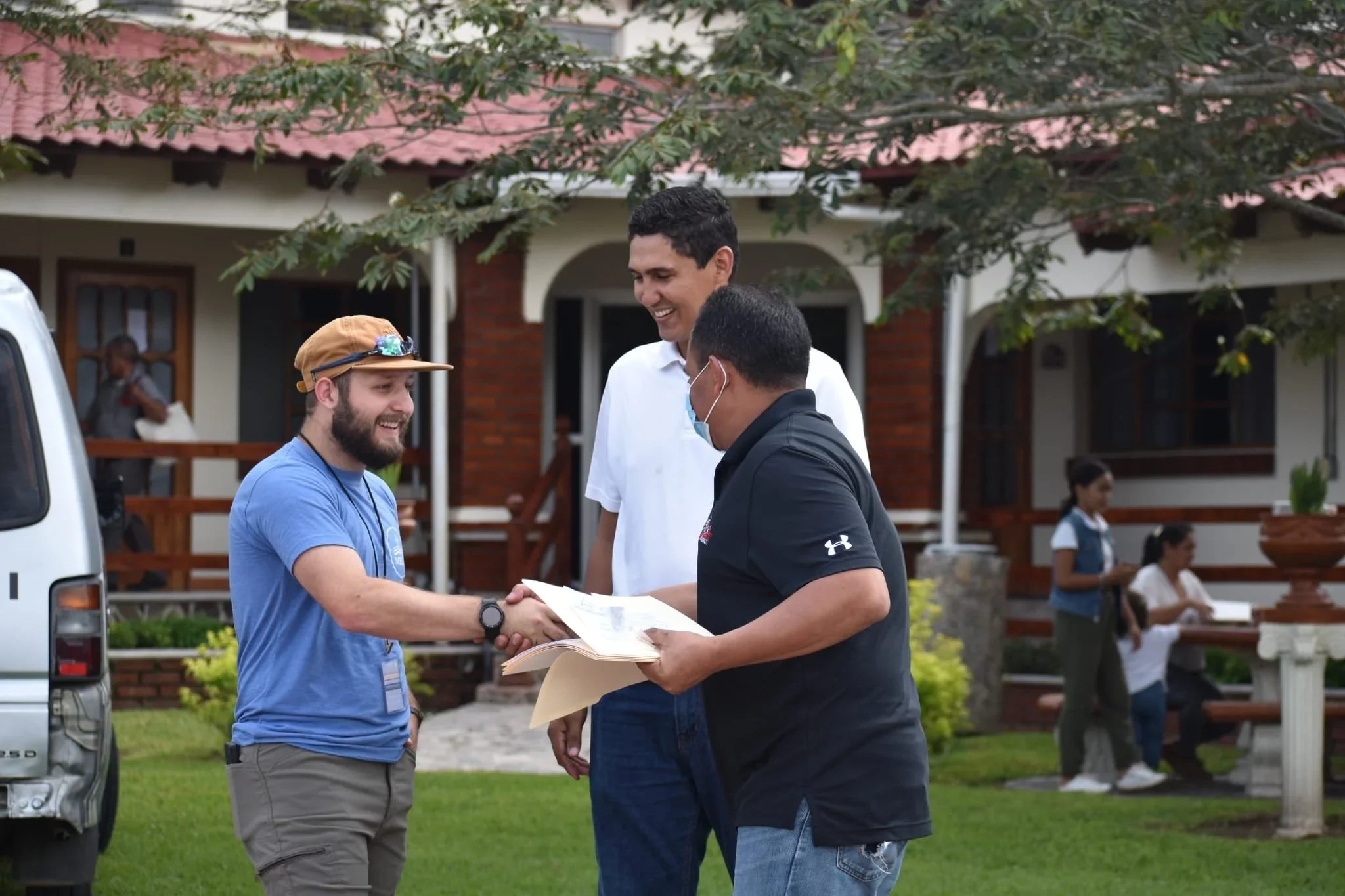 One man with a beard shaking the hands of another man who has a facemask on. There is another man with them that is smiling, taller than the rest. They are in Honduras